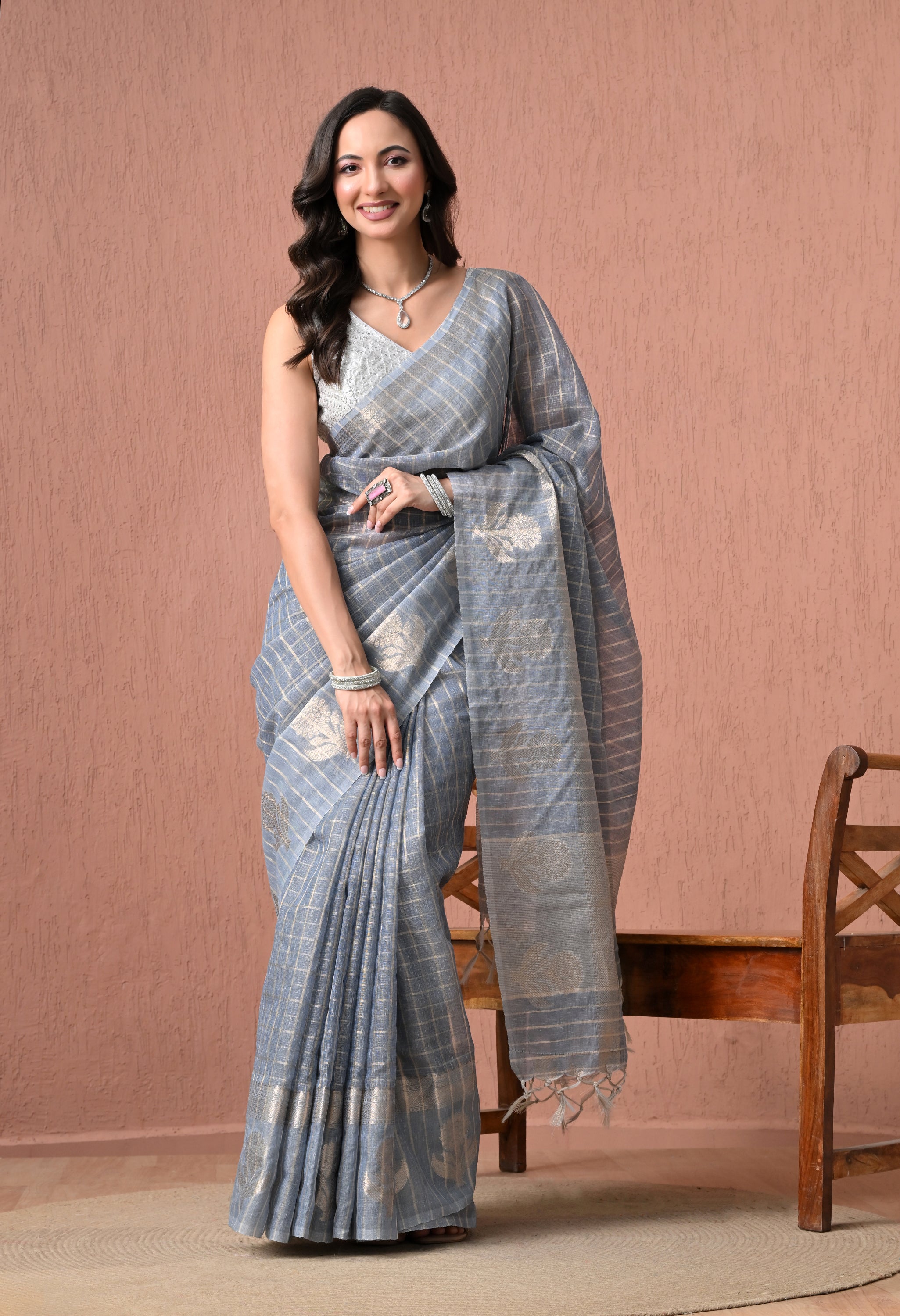 A woman smiles while posing in a steel grey cotton saree featuring silver zari checks, floral pallu motifs, and tassels, paired with a sleeveless white embroidered blouse against a textured dusty pink backdrop.
