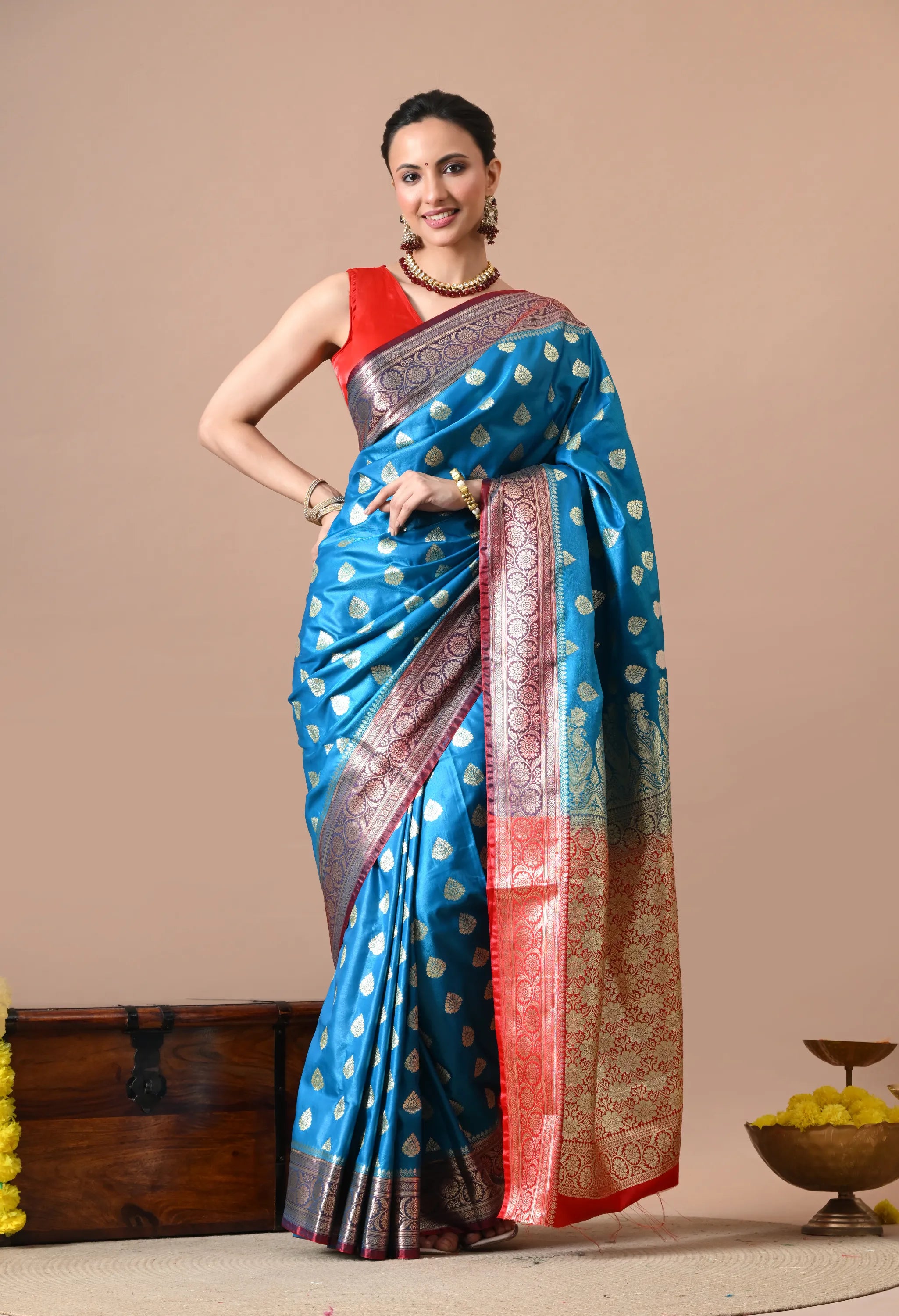 Woman posing in a teal blue silk-blend saree with gold leaf patterns and a red zari border.
