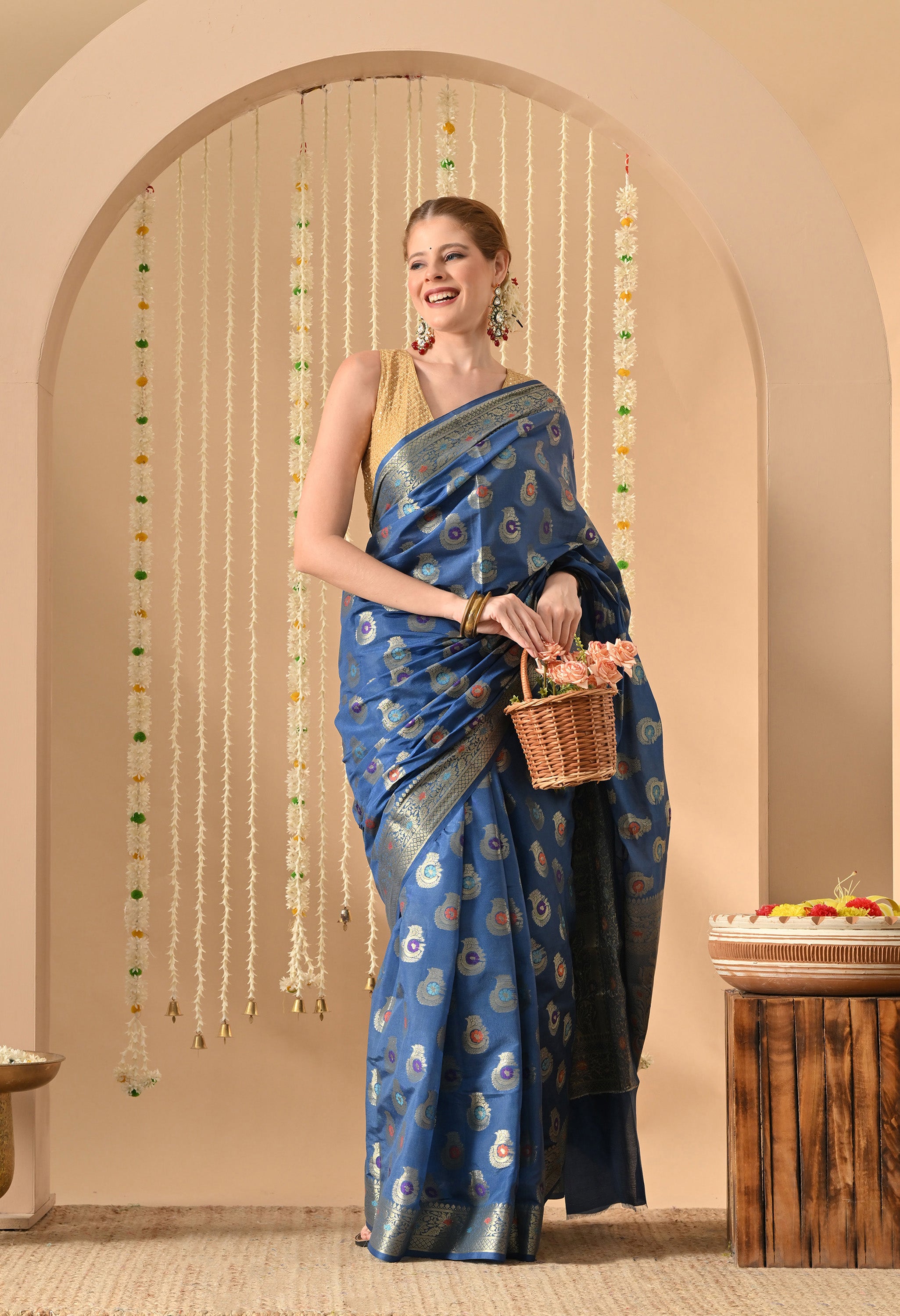 A woman models a rich blue silk saree from The Weavers Shop by Raavi. The saree features intricate Tilfi and Zari work consisting of colourful paisley and floral motifs across the body and heavy metallic gold weaving on the borders. She is styled with a sleeveless gold blouse, traditional earrings, and gold bangles. She stands in a soft peach arched alcove decorated with hanging jasmine garlands, holding a small wicker basket of pink flowers