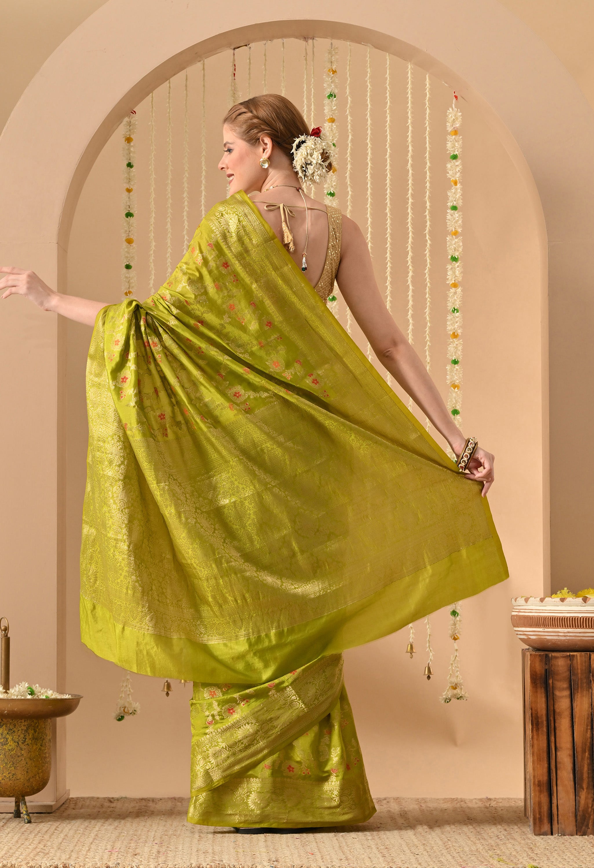 Back view of a model showcasing a luxury lime-green Banarasi silk saree from The Weavers Shop by Raavi. The image highlights the Tilfi weaving technique, featuring small floral motifs in multi-colored zari scattered across the fabric. The saree is paired with a gold sleeveless blouse with a tie-back detail and deep-neckline. The drape shows the smooth texture of the silk and the intricate gold zari work on the lower border and pallu.