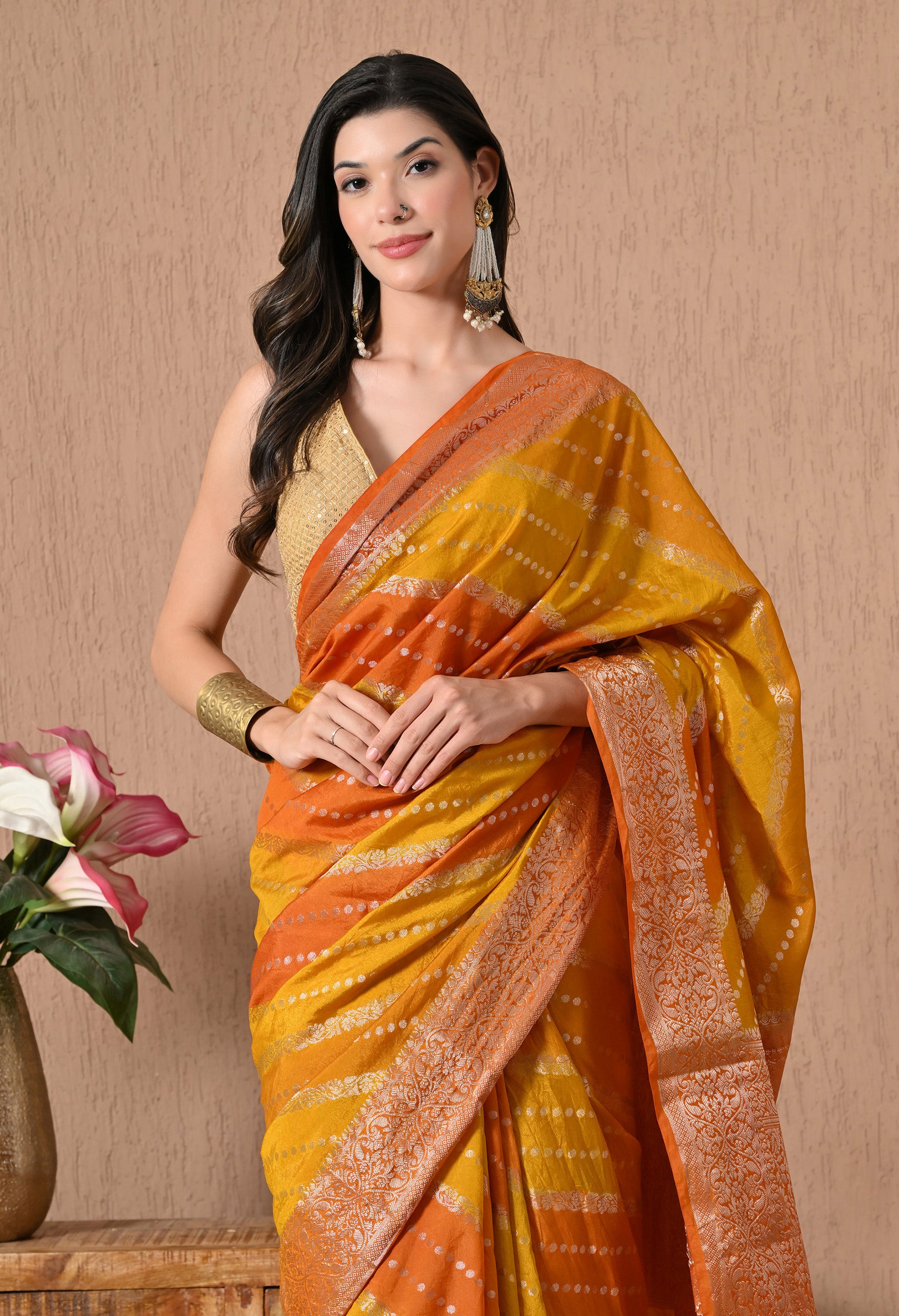 A close-up shot of a model showcasing the Rangkat Banarasi silk saree from The Weavers Shop. The image highlights the intricate golden zari work on the borders and the transition of the Rangkat diagonal stripes in mustard yellow and deep orange. Small, woven golden-toned small dots are visible across the fabric's body, and the model is styled with a sleeveless gold sequined blouse and traditional statement jewellery.
