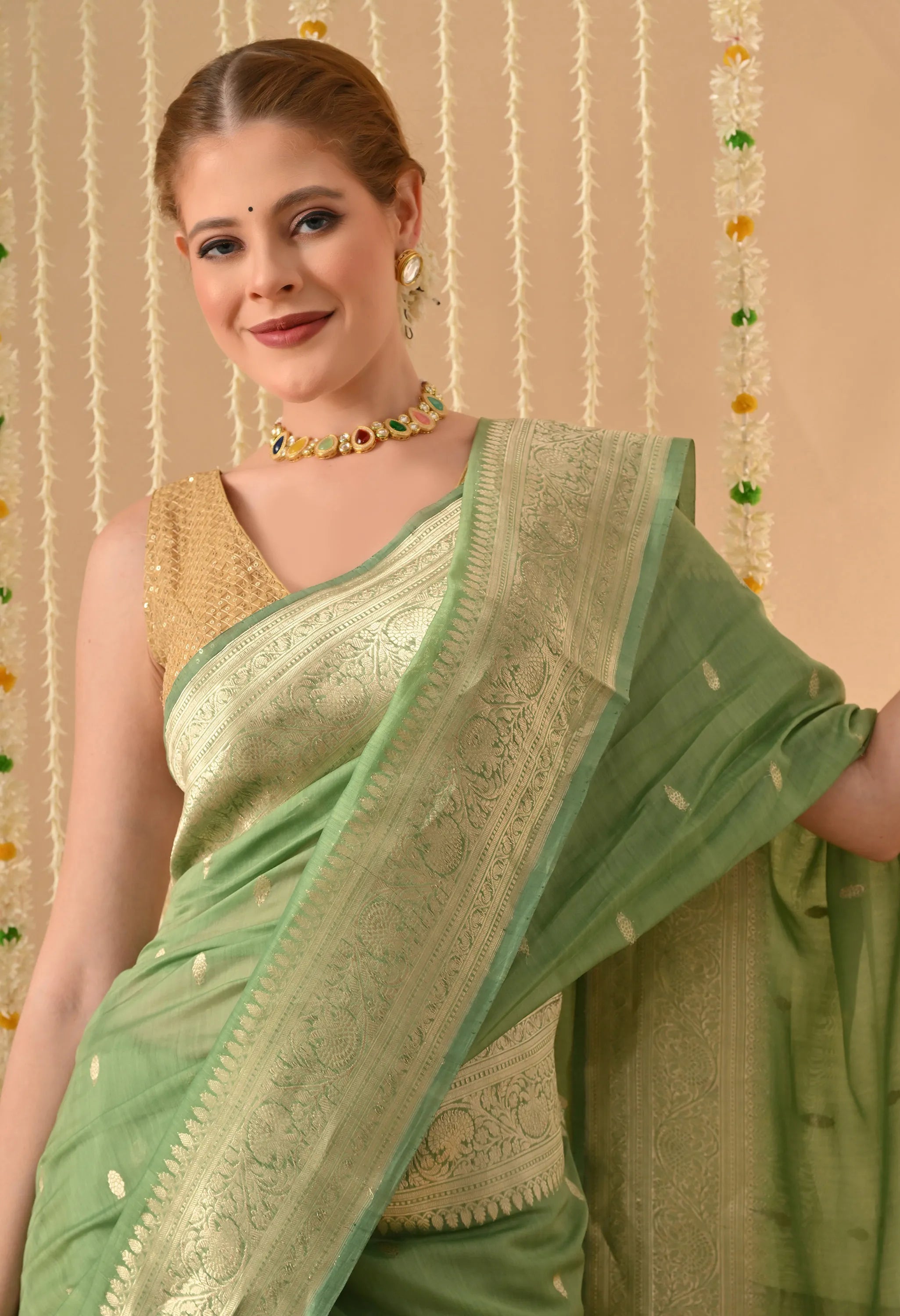 A detailed close-up shot of a woman modelling a Silk Mark Certified Mint Green Pure Silk Saree from The Weavers Shop by Raavi. The image showcases the intricate hand-woven Karuwa Weave, featuring delicate gold zari work motifs (butis) scattered across the rich, opaque katan silk fabric. The focus is on the broad, ornate gold zari border, which displays highly detailed floral and vine patterns. She is styled with a sleeveless gold blouse and a colorful stone-studded choker, set against a festive background