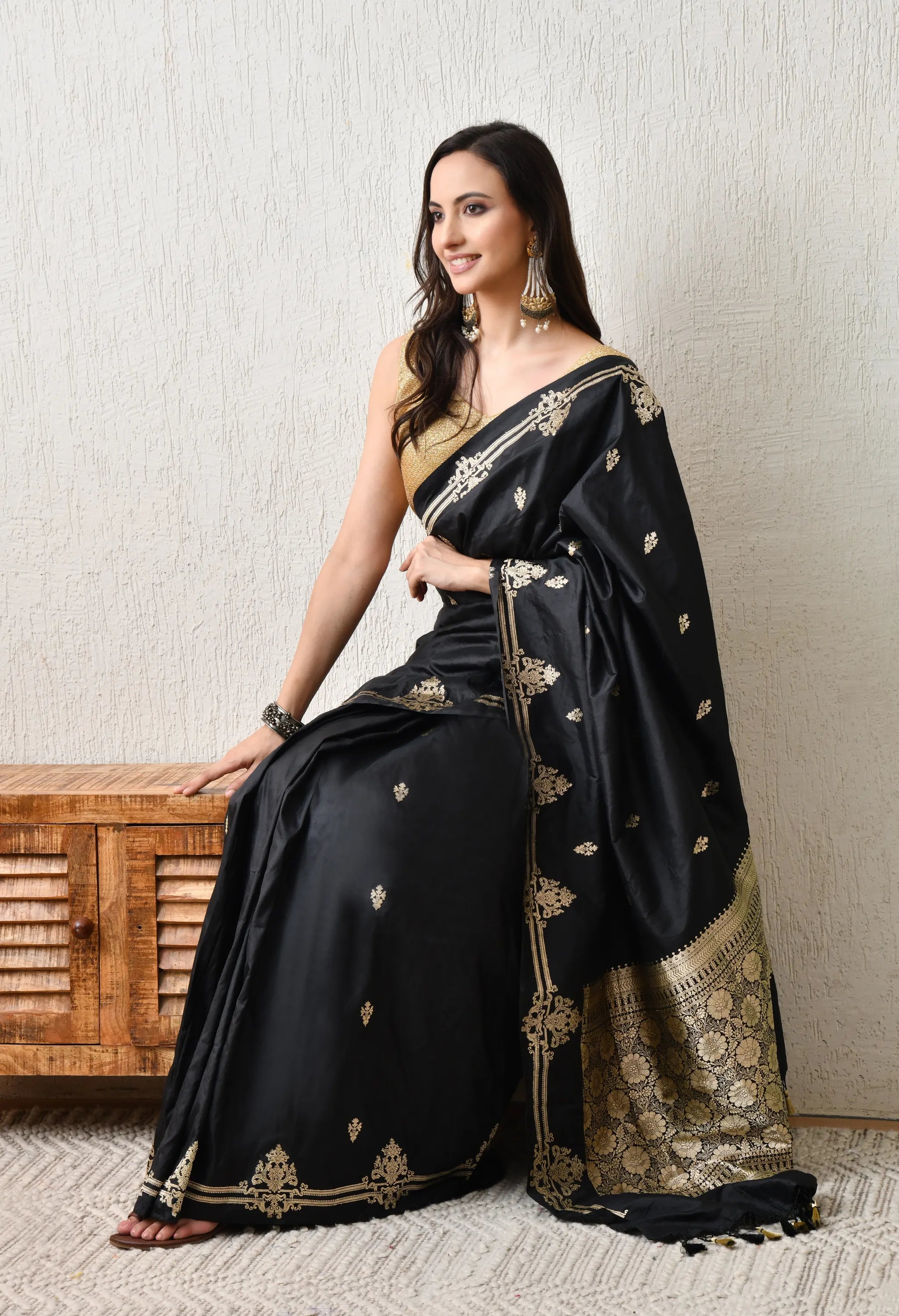 A smiling woman is seated indoors, wearing a Silk Mark Certified black silk saree from The Weavers Shop by Raavi , featuring heavy gold zari Banarasi-style work. The saree has delicate gold bootis on the body, elaborate vertical gold embroidery on the pleat edge, and a rich, wide gold brocade border and pallu. She is wearing a sleeveless gold blouse and large, tiered gold earrings, and is seated on a rustic wooden cabinet.
