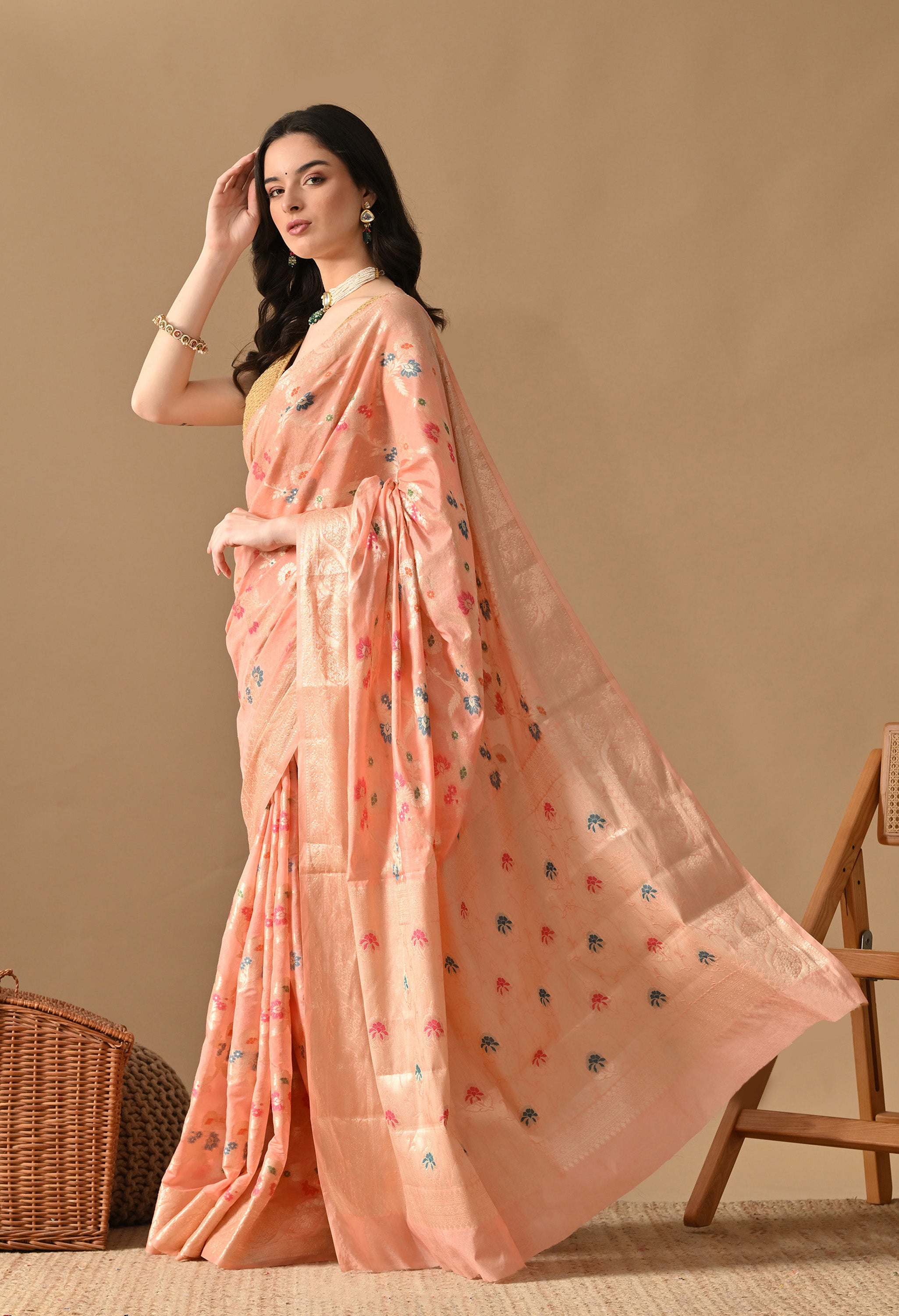 A side-profile view of a woman elegantly draped in a peach silk saree by The Weavers Shop by Raavi, highlighting the flowing pallu. The garment is decorated with Tilfi work and Zari floral motifs in pink and blue. The woman is styled with a gold sleeveless blouse and traditional jewellery, including a pearl necklace and green drop earrings. The saree is machine-woven rather than handwoven and crafted from synthetic silk.
