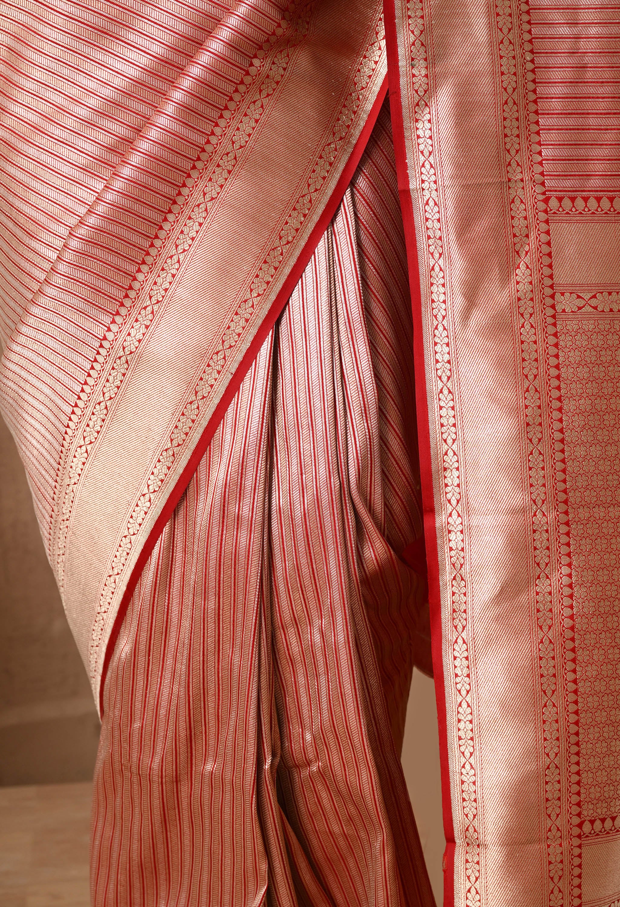 An extreme close-up shot focusing on the intricate pleats and lower drape of a Silk Mark Certified saree from The Weavers Shop by Raavi. This authentic Banarasi piece is crafted from premium Pure Katan Silk in a lustrous metallic red, gold and silver tone. The image highlights the precise hand-woven vertical zari stripes and a rich red border.