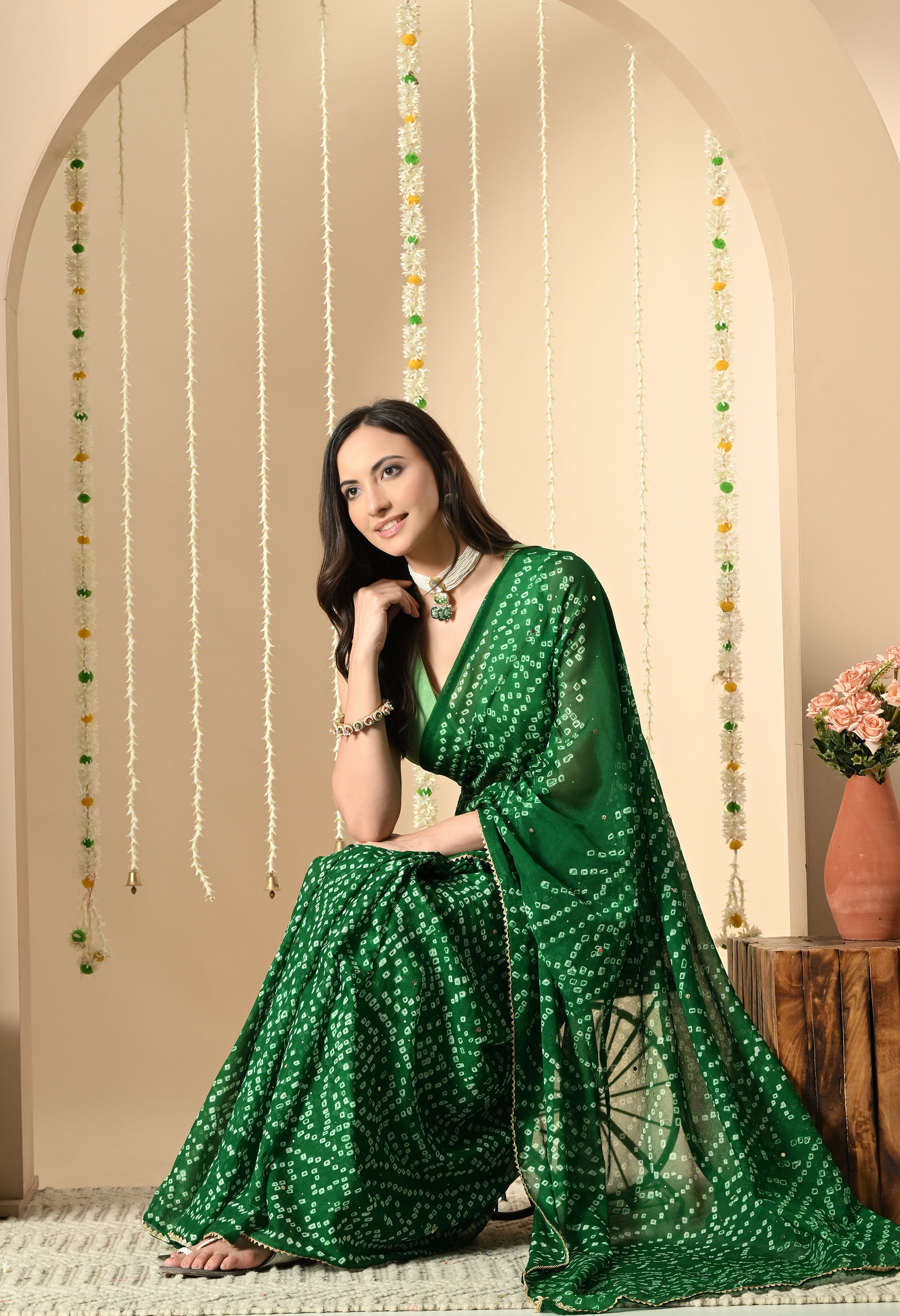 A woman is seated in a poised manner, wearing a dark green georgette sari from The Weavers Shop by Raavi, suitable for a festival or a wedding . The sari features a classic white Bandhej tie-dye design and a delicate gold trim. She is styled with a pearl choker and a sleeveless light green blouse, posed against a festive backdrop of floral decorations.