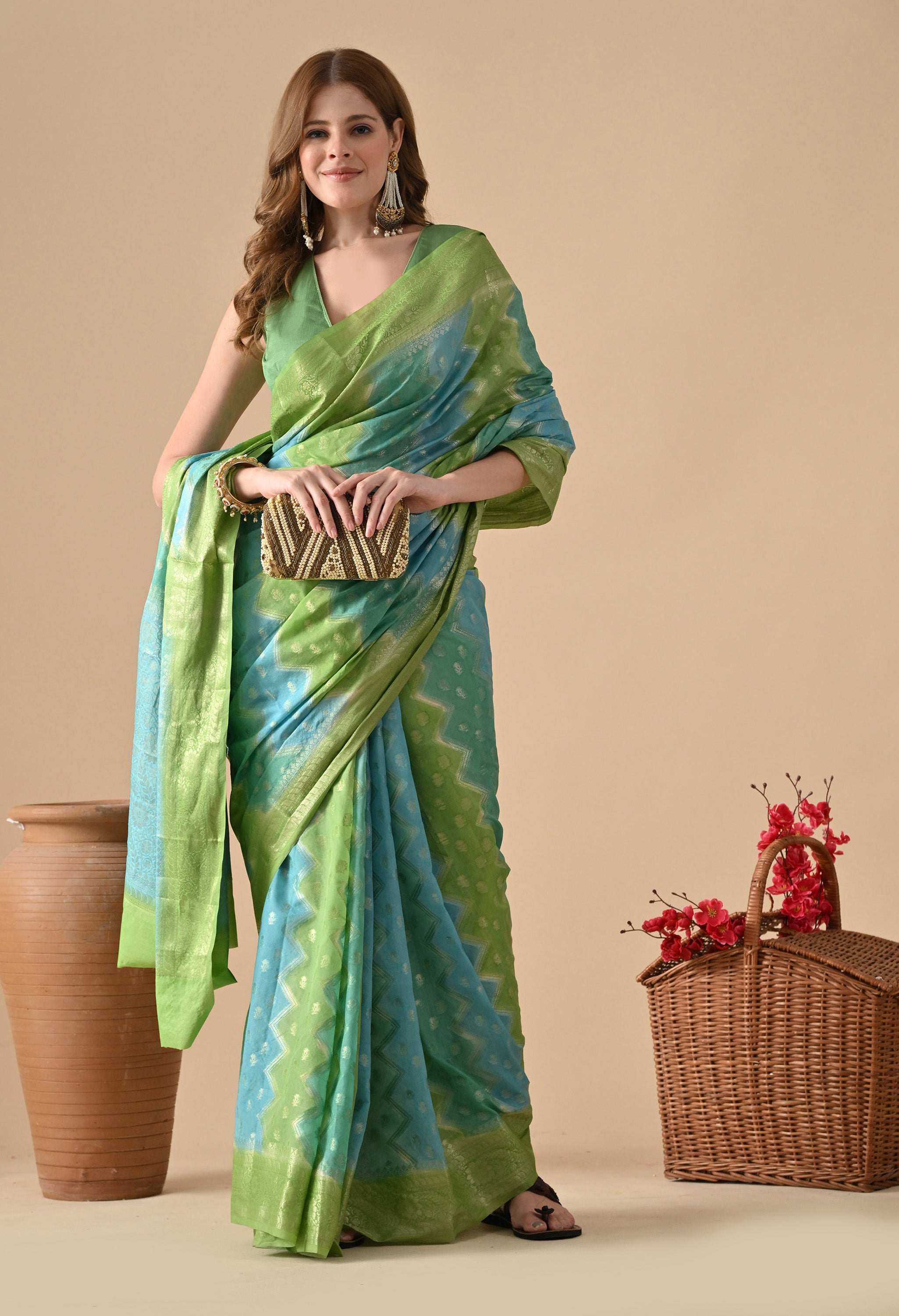 A front-facing view of a woman modeling a Bollywood-style Banarasi Rangkat saree from The Weavers Shop. She is holding a gold and brown patterned clutch bag, showcasing how to style the green and blue zigzag-patterned saree for a formal event. The saree is heavily embellished with gold zari work on the borders and body. The setting includes decorative elements like a tall ceramic pot and a wicker basket with pink flowers.