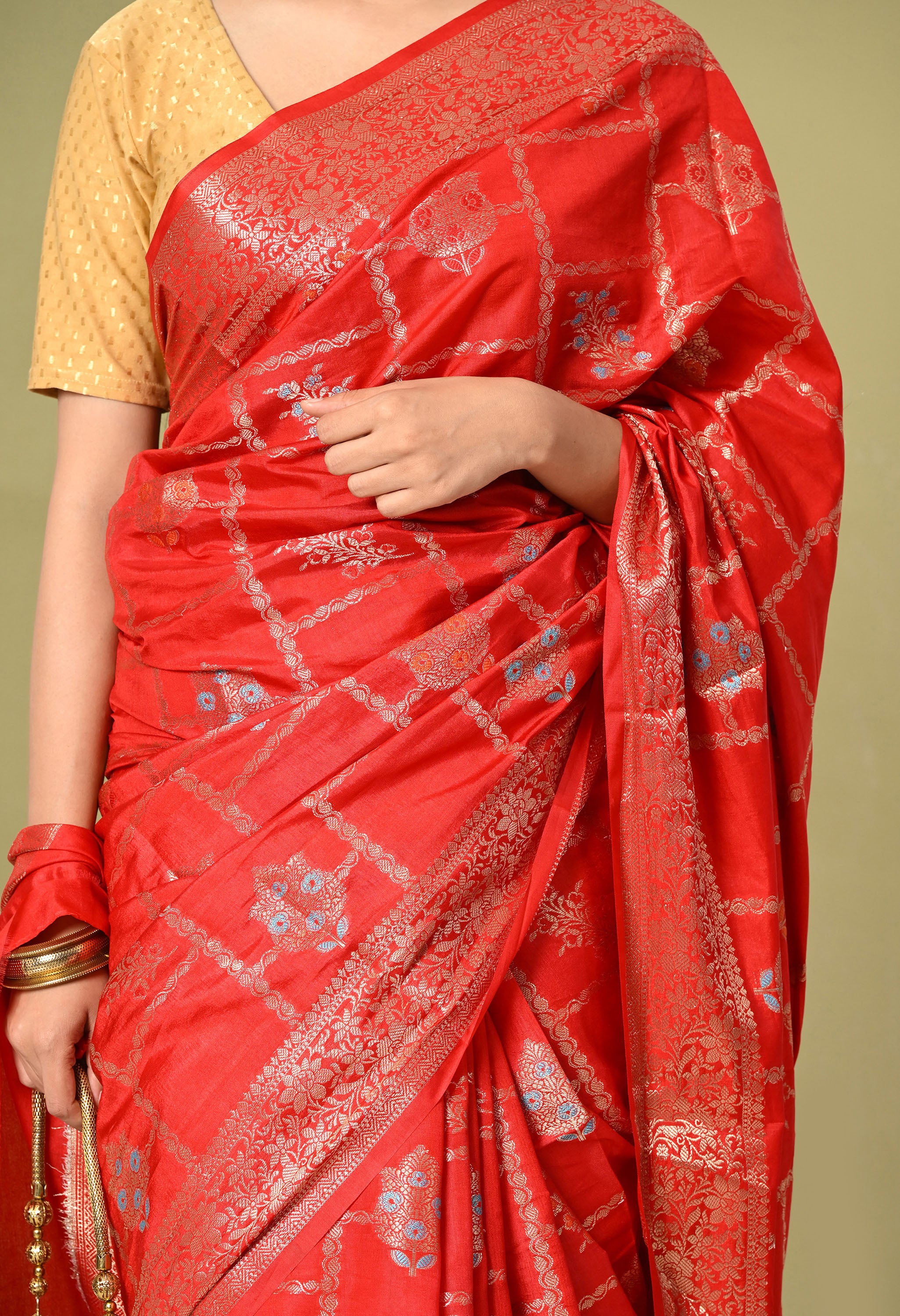 A close-up shot focusing on the rich texture and detailed Tilfi work of a saree from The Weavers Shop by Raavi. The red silk fabric features a shimmering gold zari grid and finely woven floral motifs. The shot highlights the craftsmanship of the gold border and the model's coordinated jewelry, showcasing the premium quality of The Weavers Shop by Raavi collection.