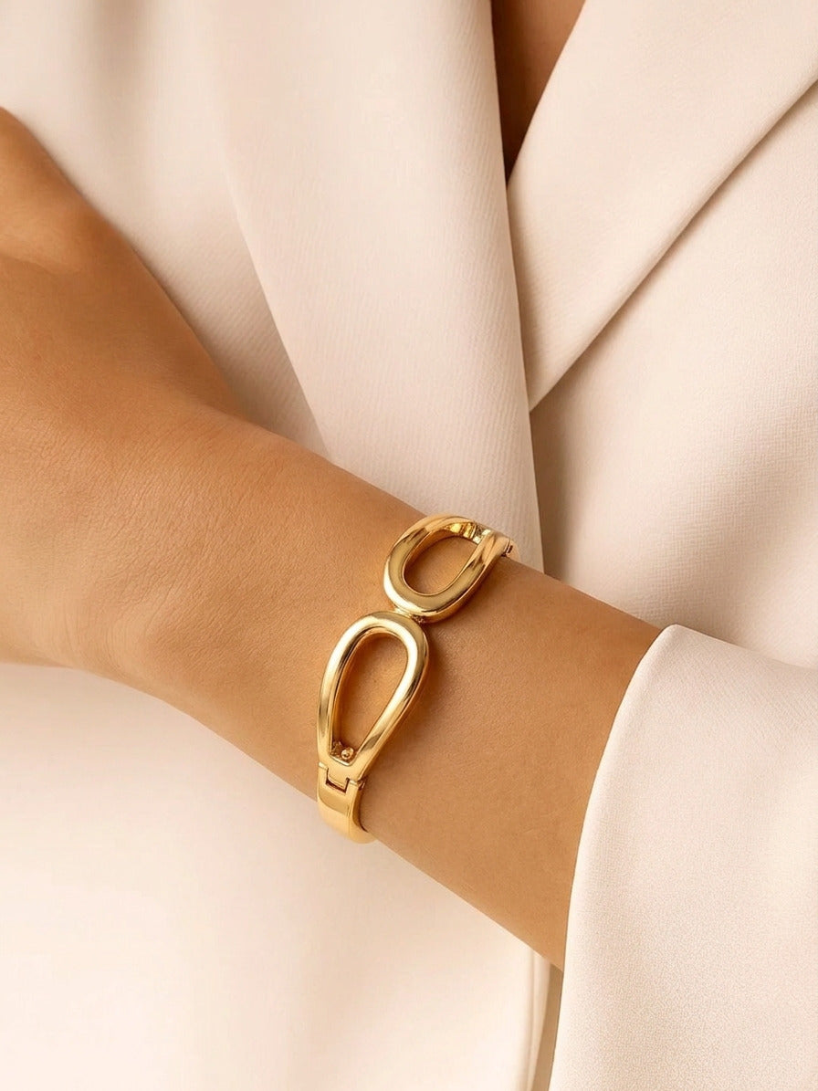 A high-polish gold hinged bangle featuring a modern double-loop geometric design on a white background.
