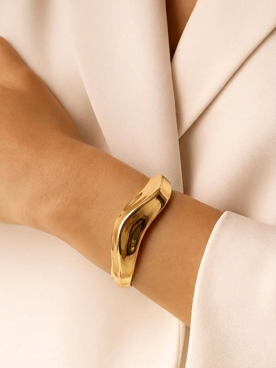 A high-shine, gold-plated sculptural bangle with an organic, wavy "liquid metal" design, shown at a 45-degree angle on a clean white background.
