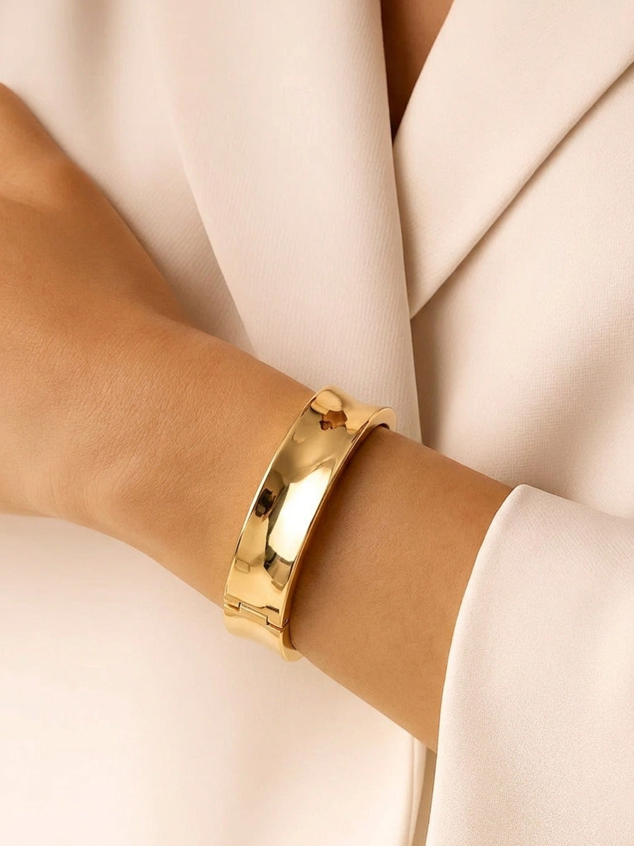 A wide, polished gold-tone hinged bangle bracelet with a sleek concave surface.
