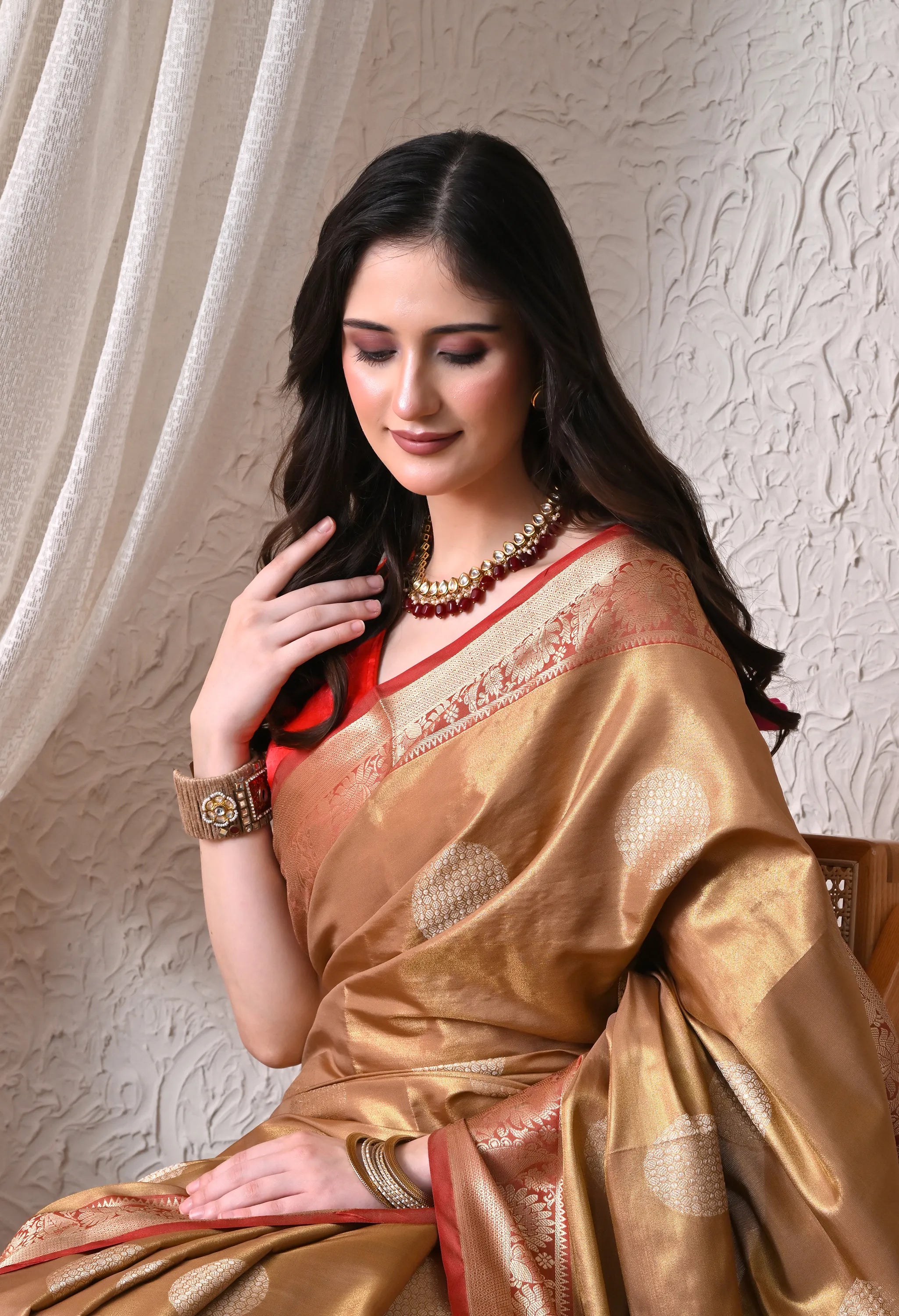 A woman elegantly carrying an antique gold silk blend saree featuring subtle circular motifs and a contrasting crimson red zari border and pallu.
