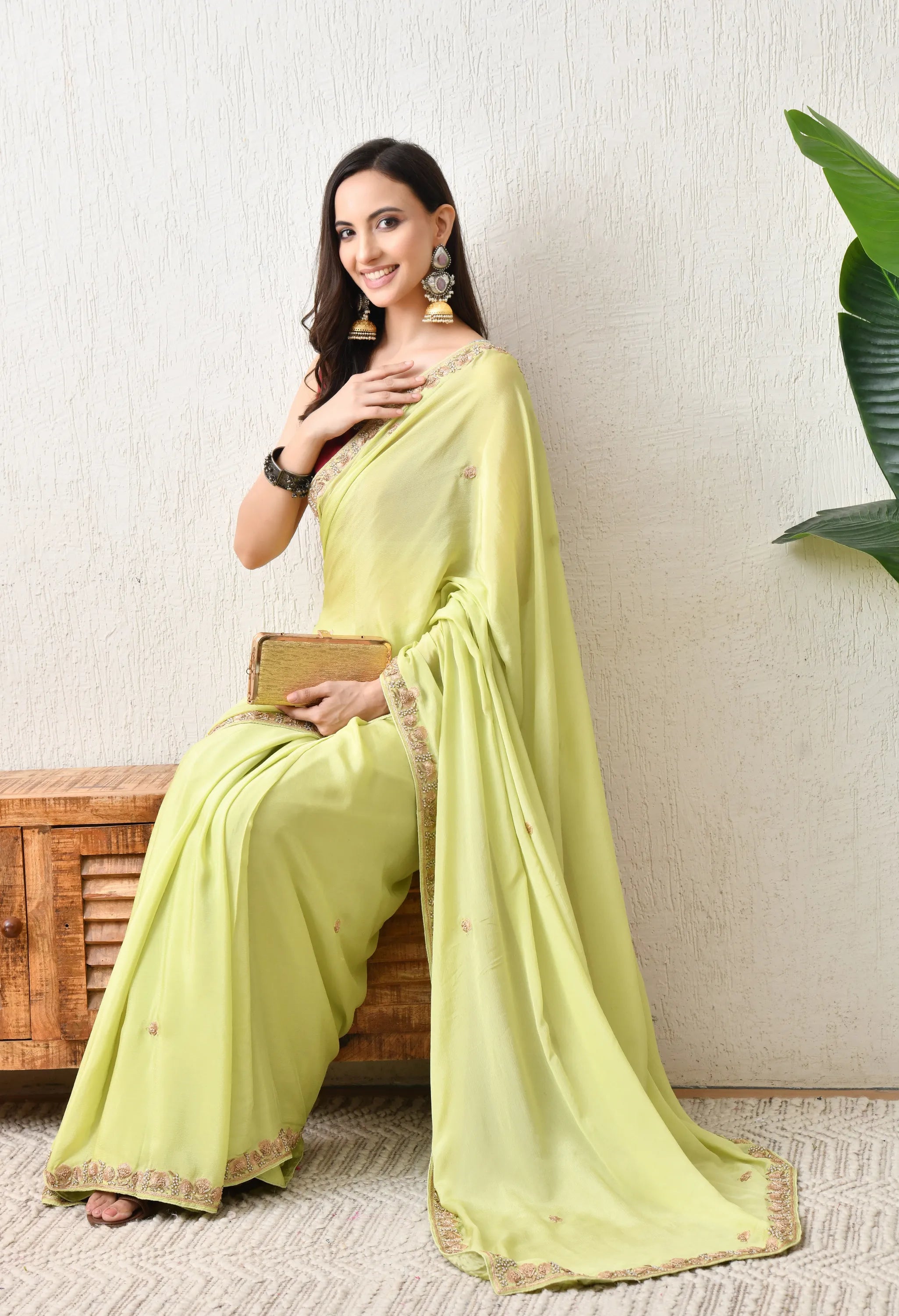 A woman sits gracefully on a rustic wooden cabinet, wearing an intricate Zardosi embroidered Pista Green chiffon saree from The Weavers Shop by Raavi. She is holding a small gold clutch bag and is accessorized with heavy traditional jhumka earrings.
