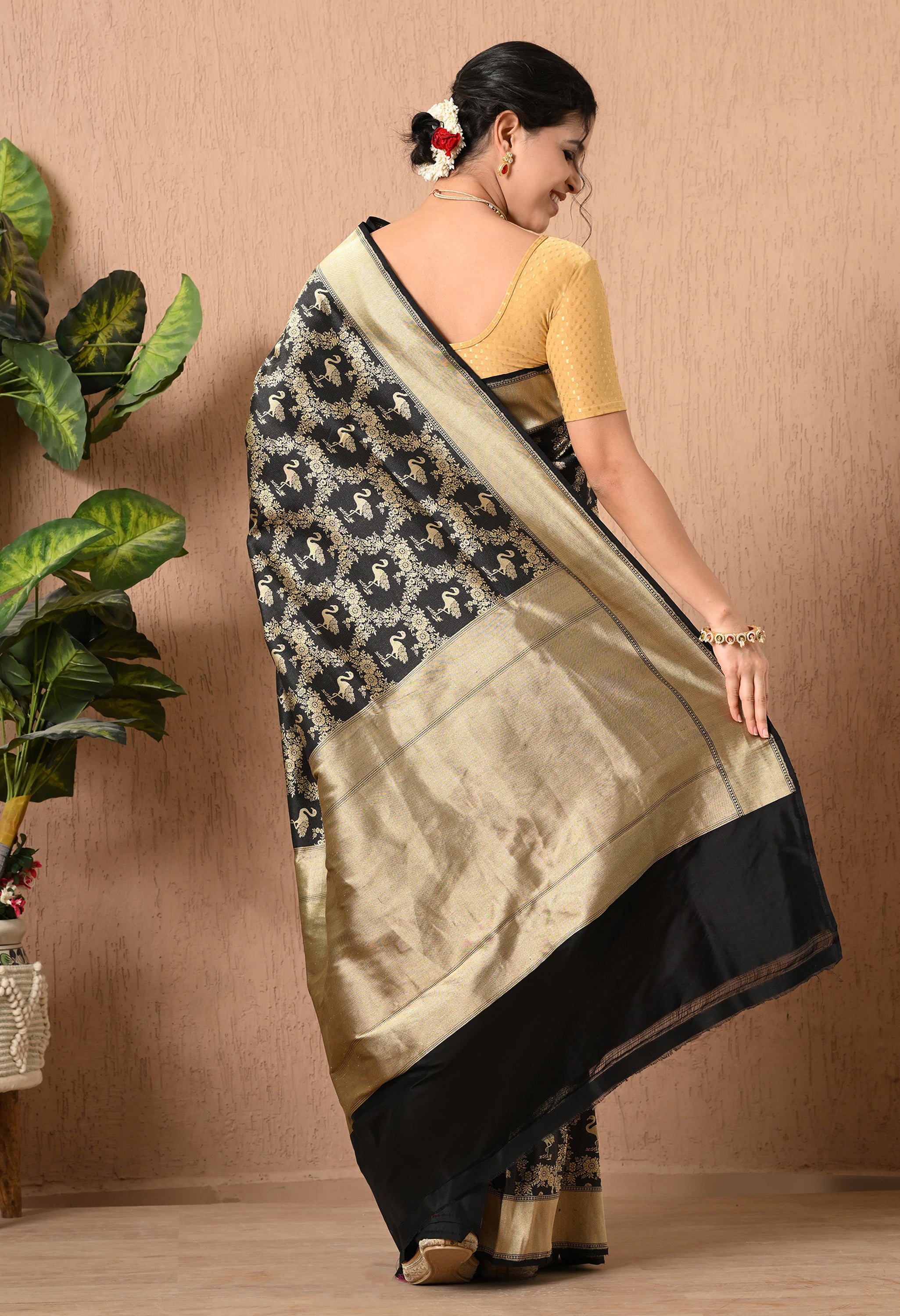 A back view of a woman showcasing a sophisticated black handd-woven pure Katan Silk saree from The Weavers Shop by Raavi. The fabric features an intricate hand-woven zari Shikarga design, illustrating traditional motifs of animals and vegetation in shimmering gold. This Silk Mark Certified piece is finished with a solid black border at the hem and a wide, textured gold pallu. She is wearing a gold blouse, and her hair is styled in a bun.