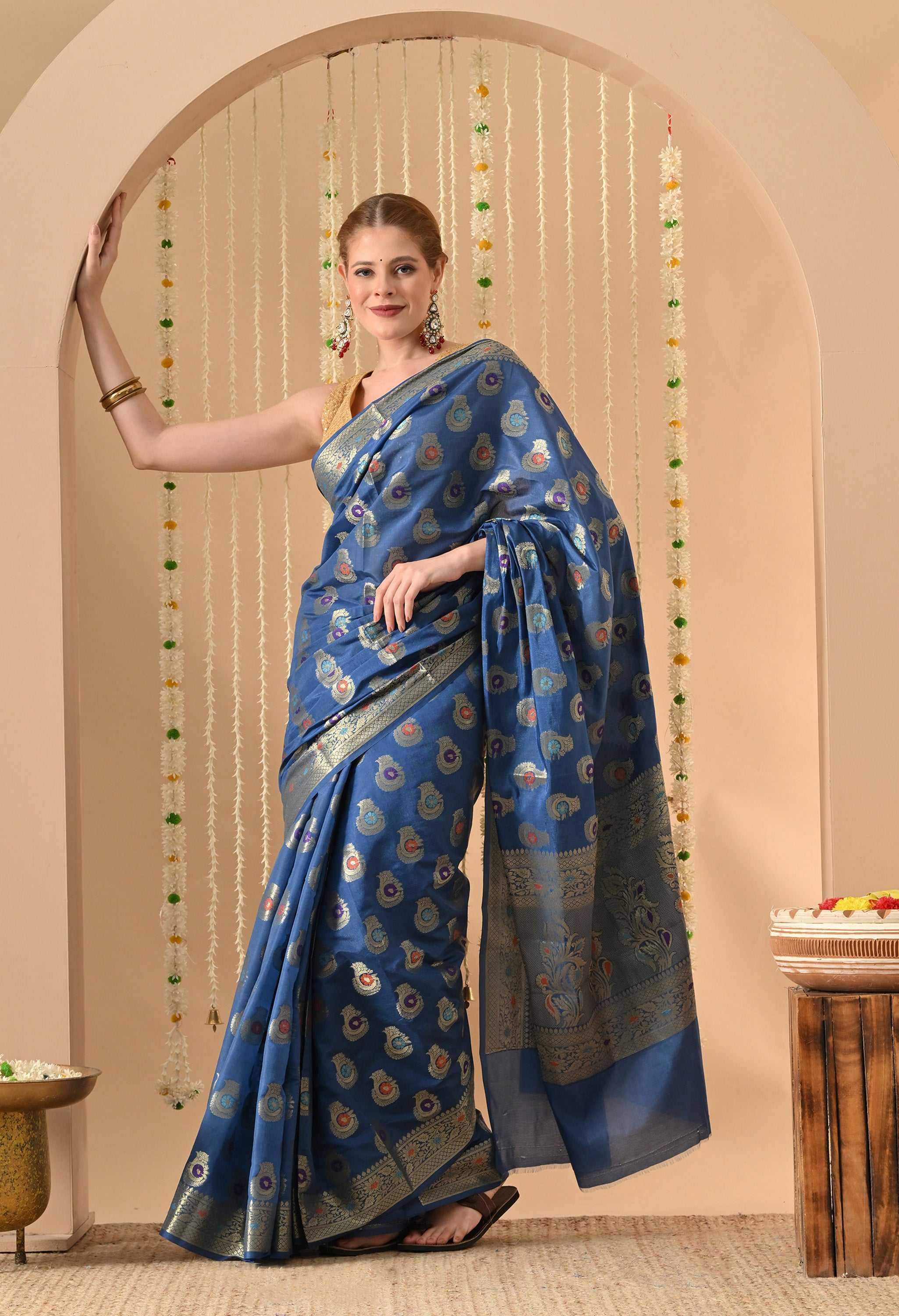 A woman smiling and posing in a blue Banarasi-style silk saree by The Weavers Shop by Raavi. The saree features intricate Tilfi work with metallic zari in traditional paisley motifs across the body and a decorative border. She is wearing a sleeveless gold blouse and statement earrings, standing against a neutral background with floral hangings.