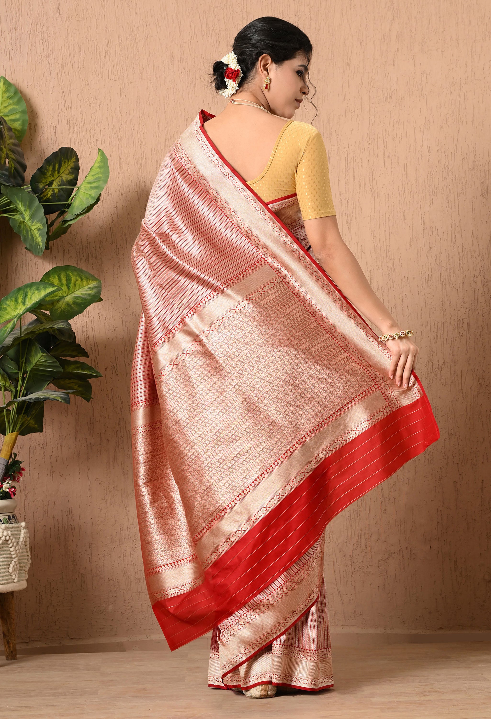 A back view of a woman showcasing an elegant Silk Mark Certified saree from The Weavers Shop by Raavi. The garment is crafted from premium Pure Katan Silk in a shimmering metallic red, gold and silver tone, featuring a pattern of hand-woven vertical zari stripes across the body. This authentic Banarasi piece is defined by a broad, intricate gold zari border and a heavily detailed pallu, finished with a solid red border at the hem. She is wearing a mustard-gold textured blouse and a floral hair arrangement.