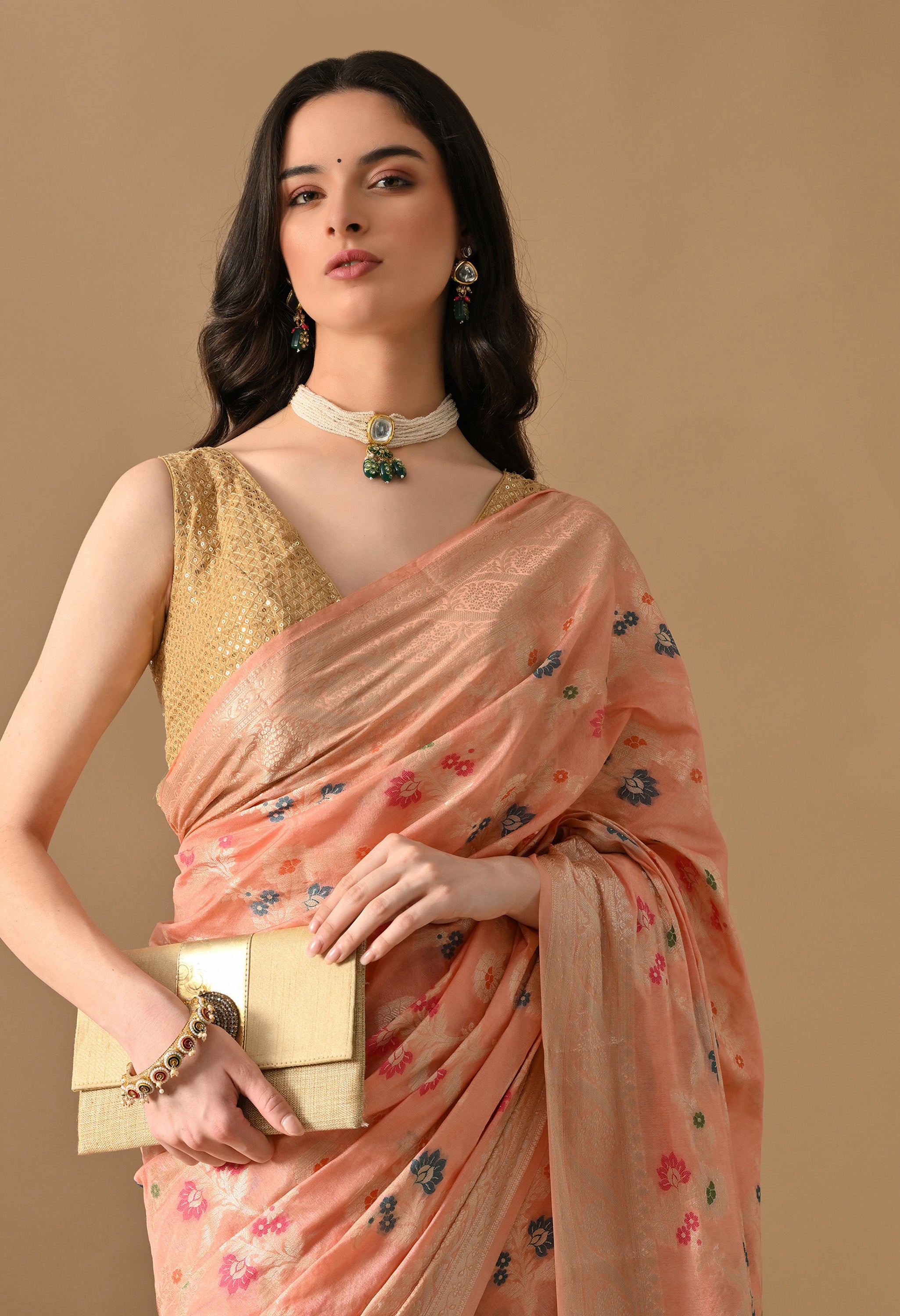 A close-up shot focusing on the upper half of a woman wearing a peach silk saree by The Weavers Shop by Raavi. The image highlights the fine Tilfi work with Zari floral details on the woven fabric. She is styled with a gold sequined blouse, a gold clutch, and a multi-strand pearl choker.