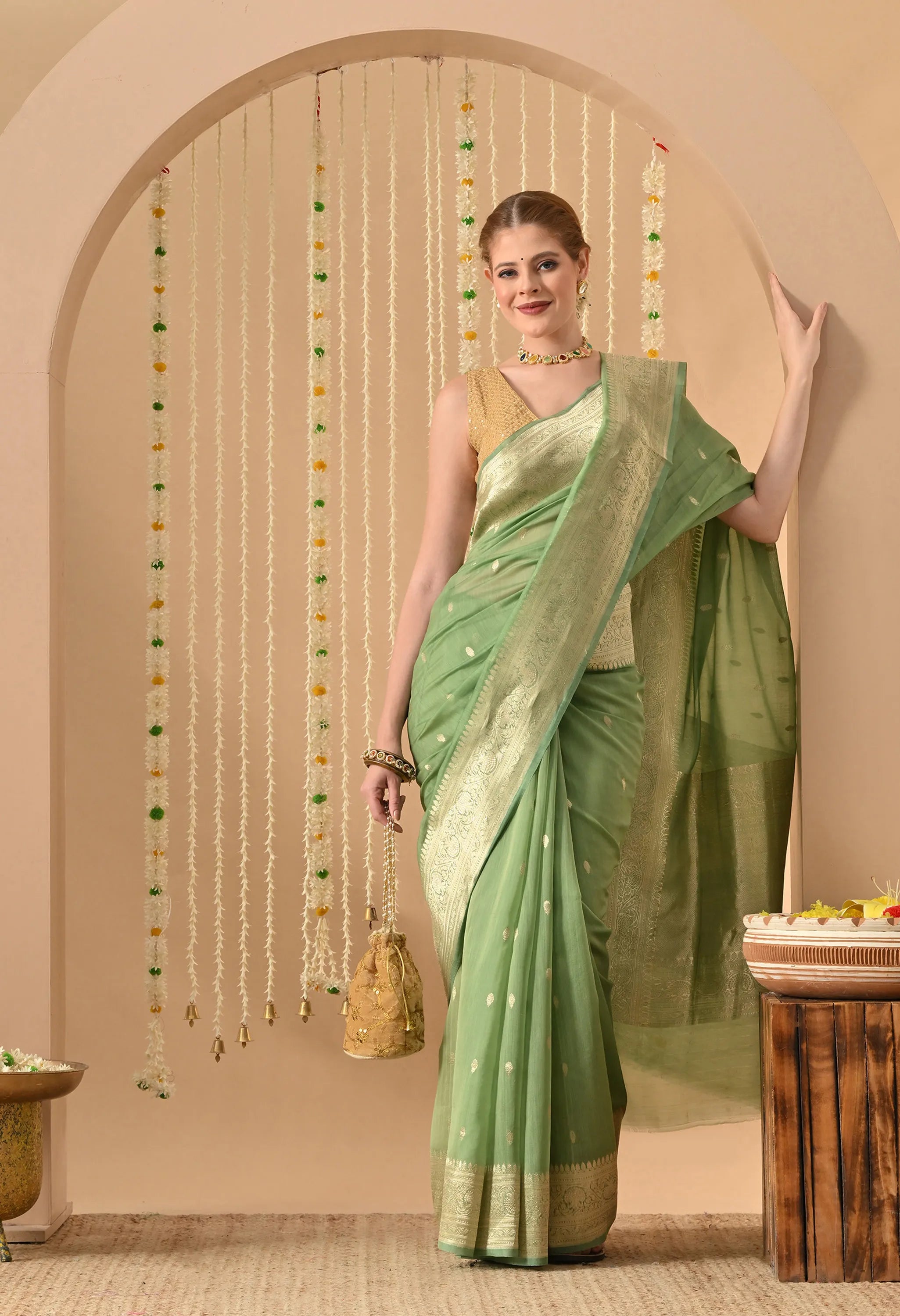 A woman models a Silk Mark Certified Mint Green Pure Silk Saree from The Weavers Shop by Raavi. This hand-woven Banarasi garment features the traditional Karuwa Weave, showcasing delicate gold zari work butis (motifs) across a rich, opaque Katan silk base. The saree is designed with a broad, shimmering gold zari border and a matching pallu, paired with a sleeveless gold tblouse. The festive setting, decorated with hanging jasmine garlands, highlights the soft, regal glow of the pastel green silk.