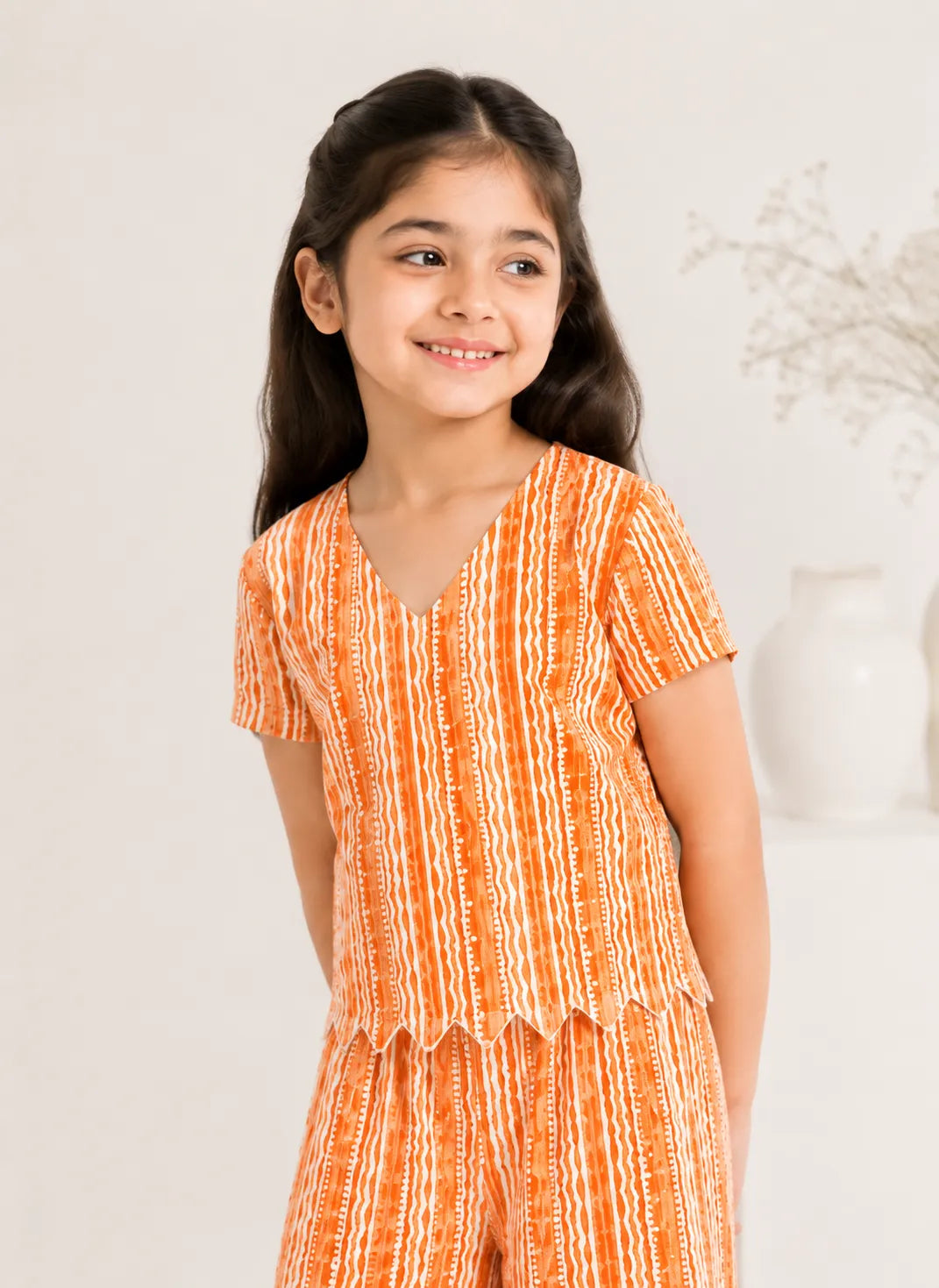 Young girl smiling in an orange and white printed two-piece co-ord set featuring a scallop-hem top and wide-leg trousers.
