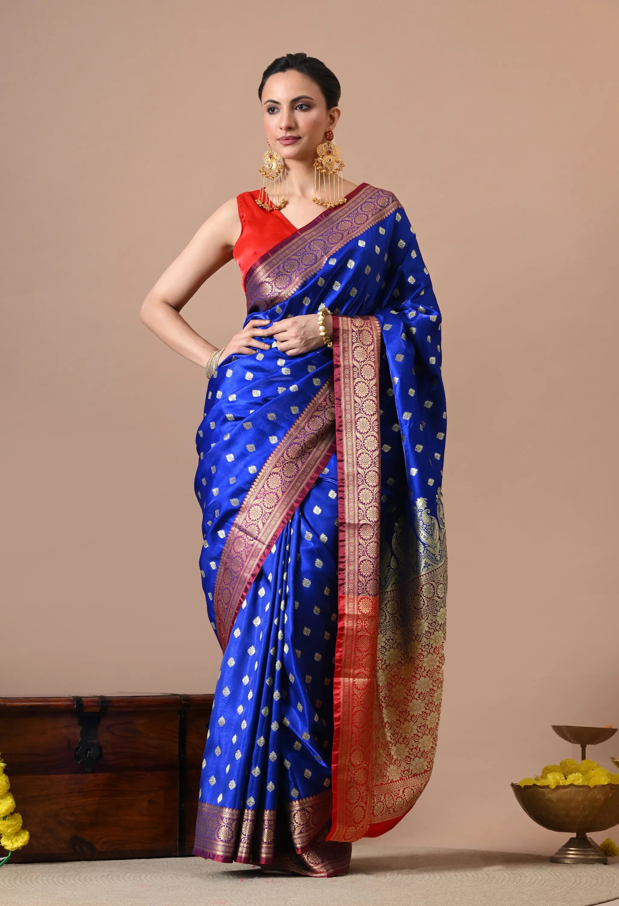 A woman posing in a royal blue Banarasi silk saree featuring small gold-toned woven motifs (Butis) across the body and a contrasting red and gold zari border and pallu.