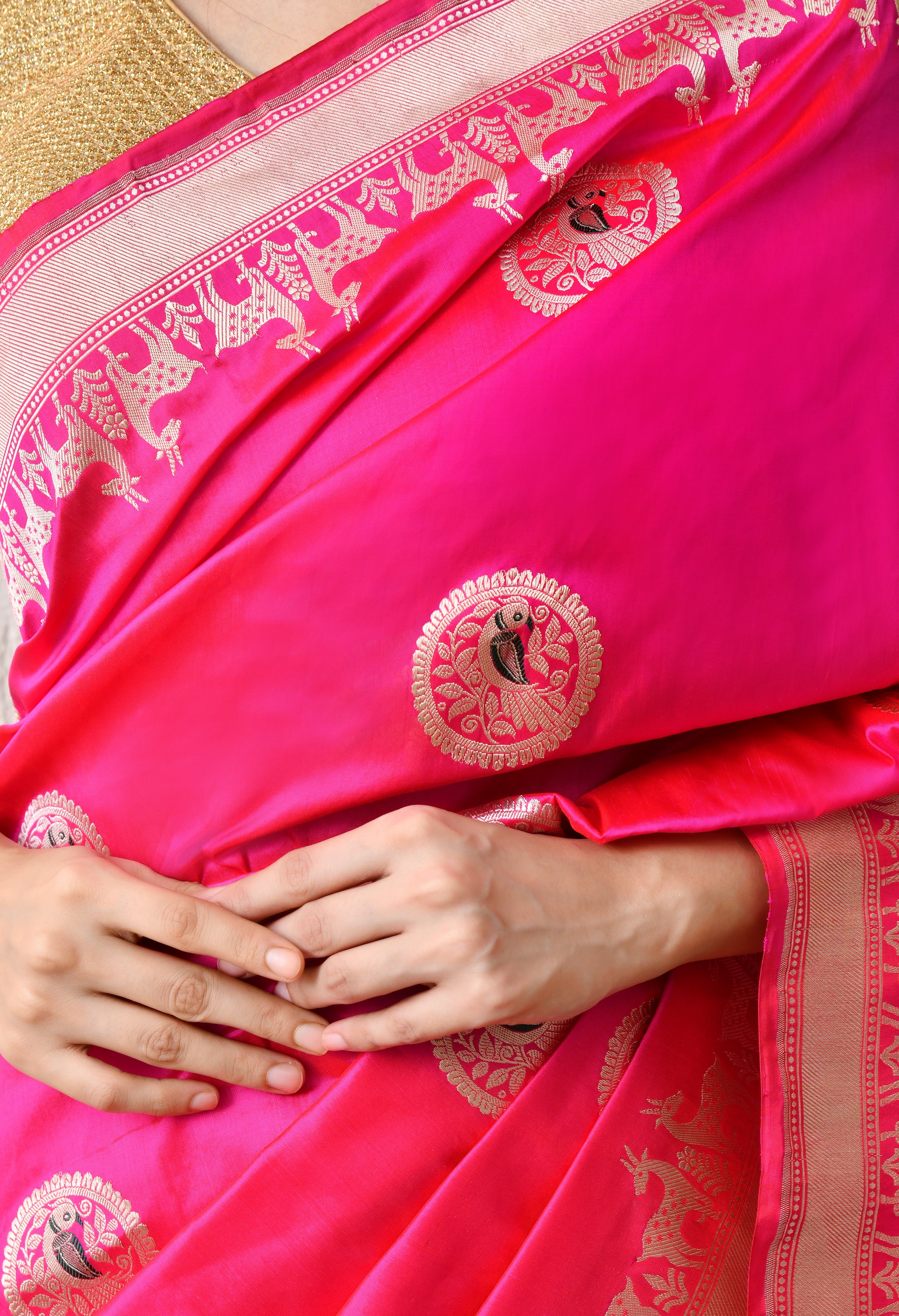 An extreme close-up shot focusing on the golden zari craftsmanship of the Hand Woven Katan Silk saree. The image highlights the intricate bird design within a circular buti and the fine detail of the deer motifs on the Shikarga border. The rich, hot pink texture of the Silk Mark Certified fabric and the gold weave of the accompanying blouse are clearly visible.