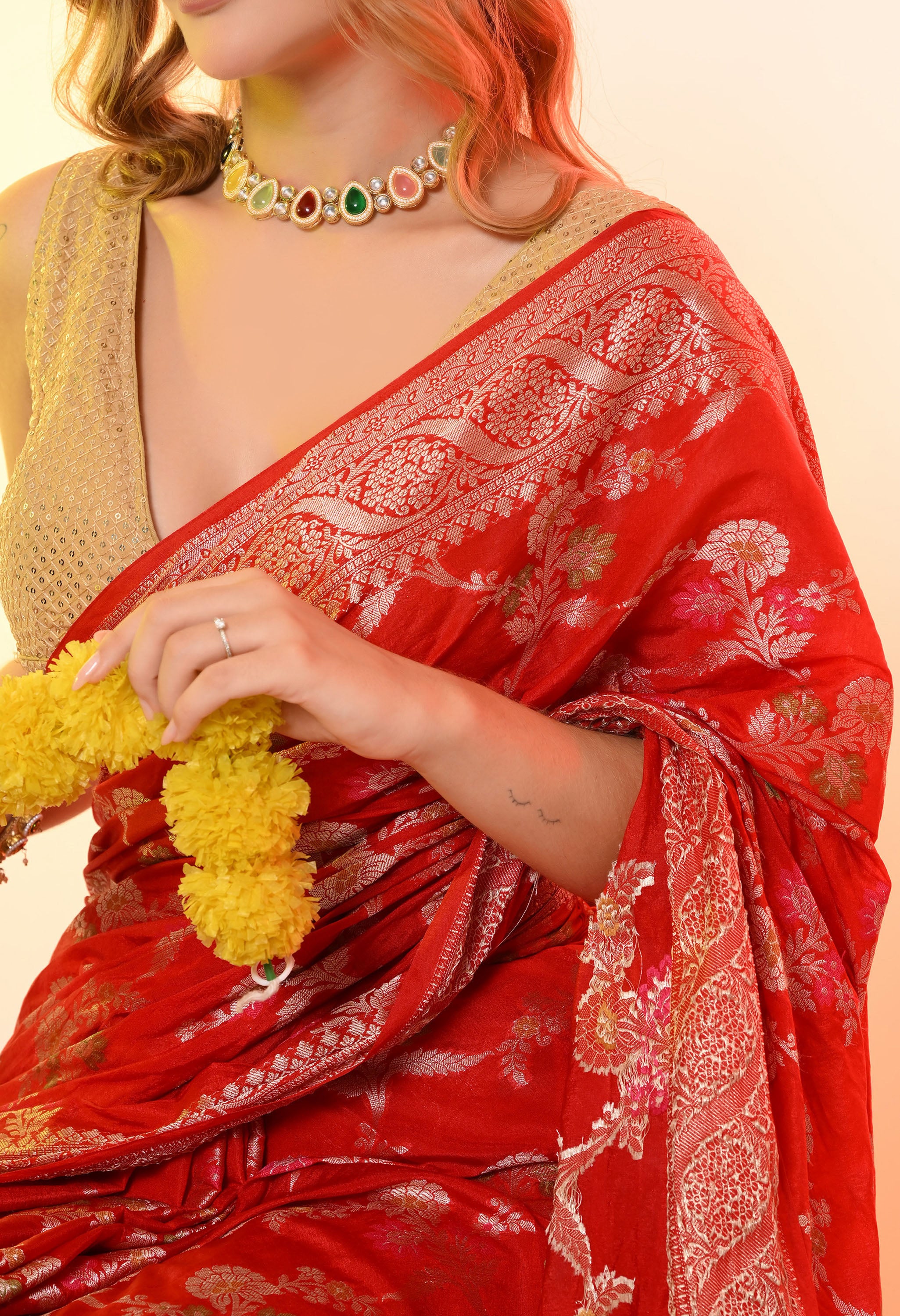 A close-up detail shot of the red Banarasi  silk- saree by The Weavers Shop by Raavi, highlighting the golden zari border and colourful floral Tilfi work. The image shows the woman’s hand holding a yellow flower garland, wearing a kundan choker with multi-colored stones.