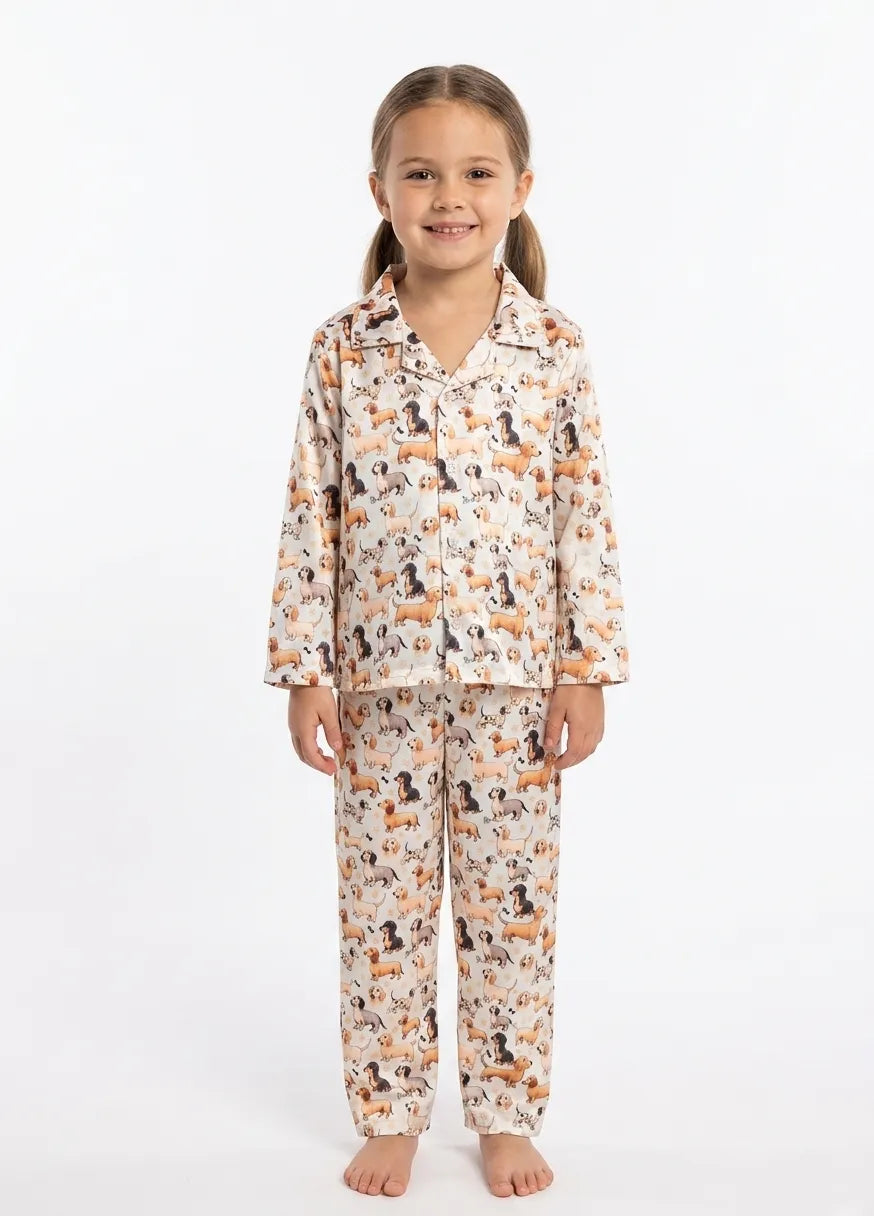 A young girls standing against a plain white background wearing a cream-colored satin pajama set with an all-over print of dachshund dogs.