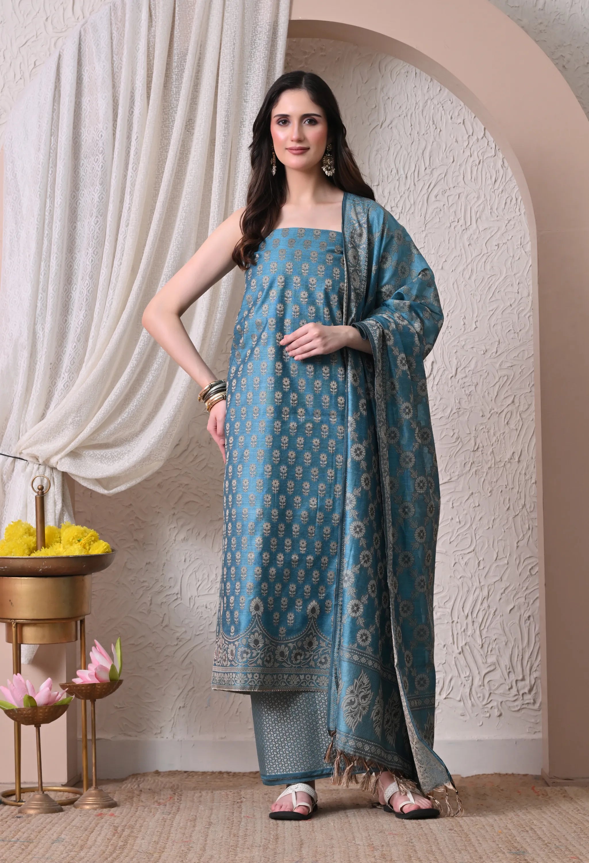 A woman posing in a luxurious teal blue and gold woven silk salwar suit set featuring detailed floral motifs, a traditional border design, and a matching tasseled dupatta.
