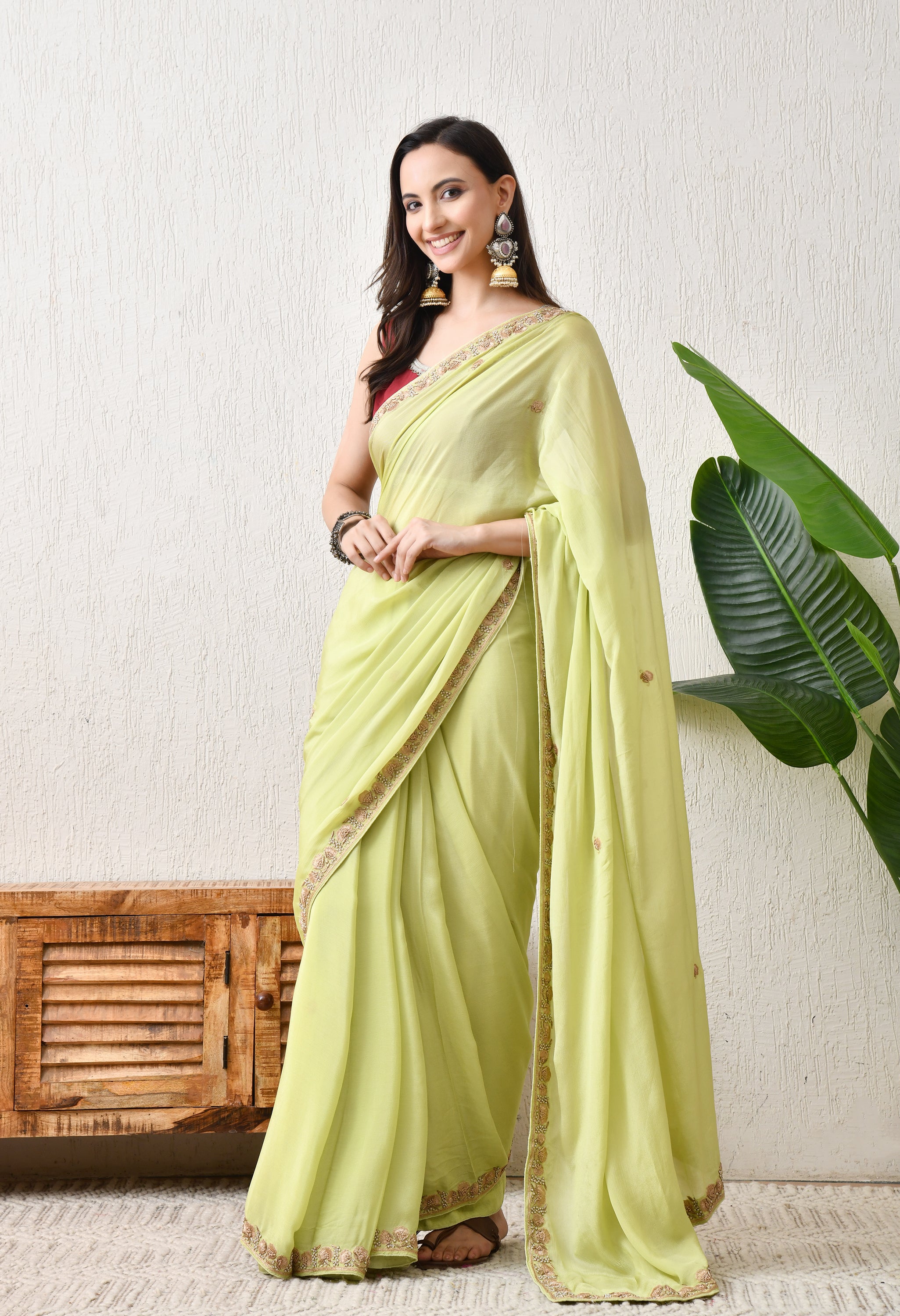 A woman stands in a 3/4 full-length pose wearing a Pista Green chiffon saree from The Weavers Shop by Raavi. The saree features a golden Zardosi hand-worked border and is paired with a maroon sleeveless blouse. She is accessorized with large traditional jhumka earrings and a dark bangle, standing in a room with a rustic wooden cabinet and a large green indoor plant.