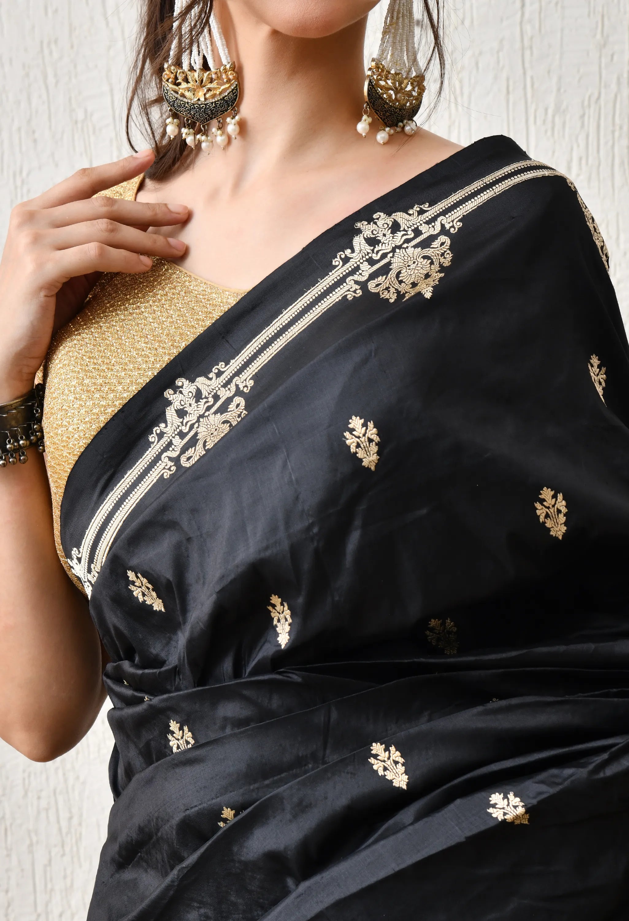 A close-up of a woman showcasing a Silk Mark Certified black Katan silk saree from The Weavers Shop by Raavi. The saree has intricate gold zari embroidery, featuring large, stylised gold motifs on the shoulder pleats and a thin gold border. She is wearing a metallic gold sleeveless blouse, a stack of black bangles on her left wrist, and large, tiered gold and white beaded earrings while lightly touching her neck.