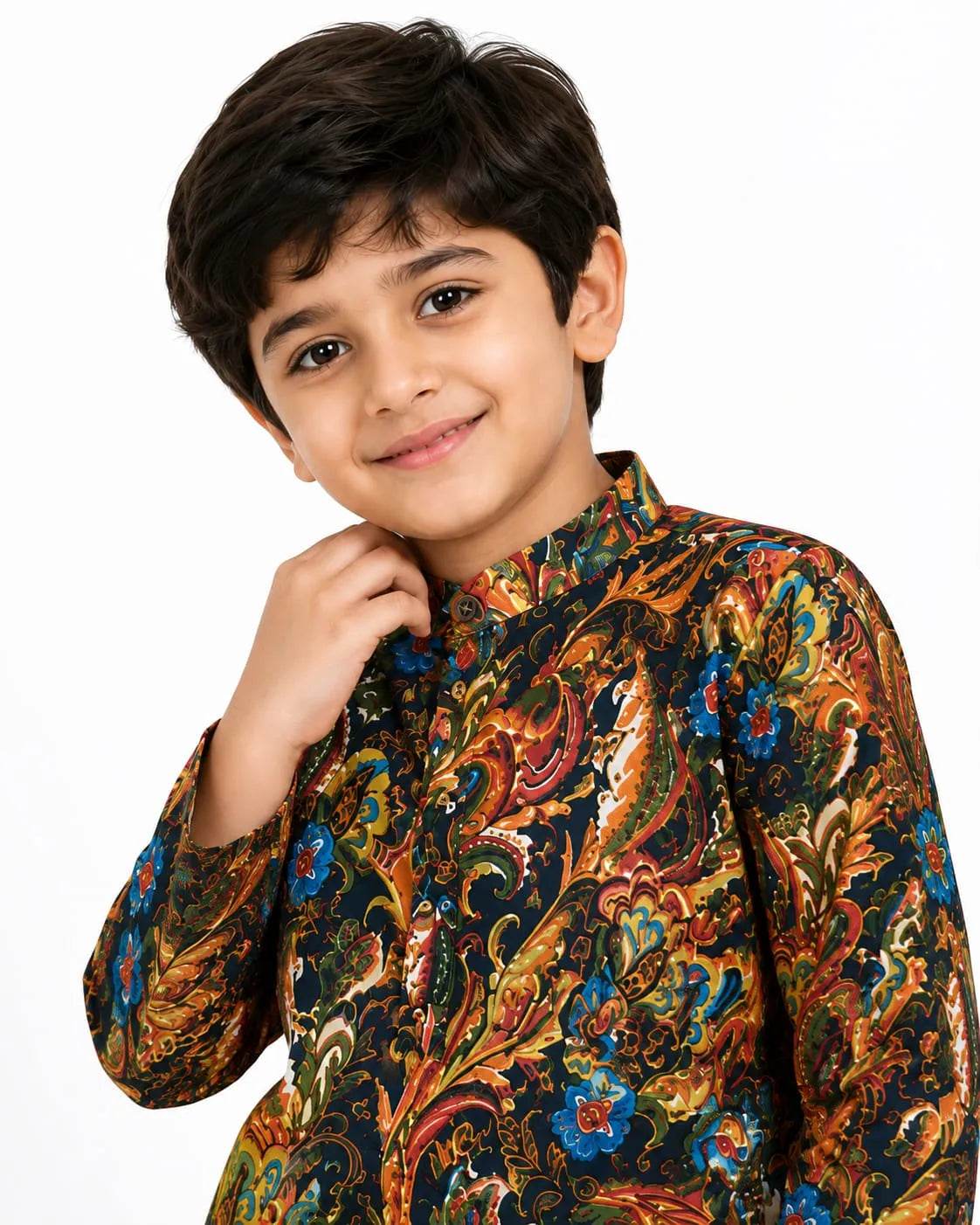 A young boy standing against a plain white studio background, wearing a multi-colored Kalamkari printed long-sleeve cotton kurta in shades of navy blue, orange, red, and green, paired with white pyjama trousers and brown textured ethnic juttis
