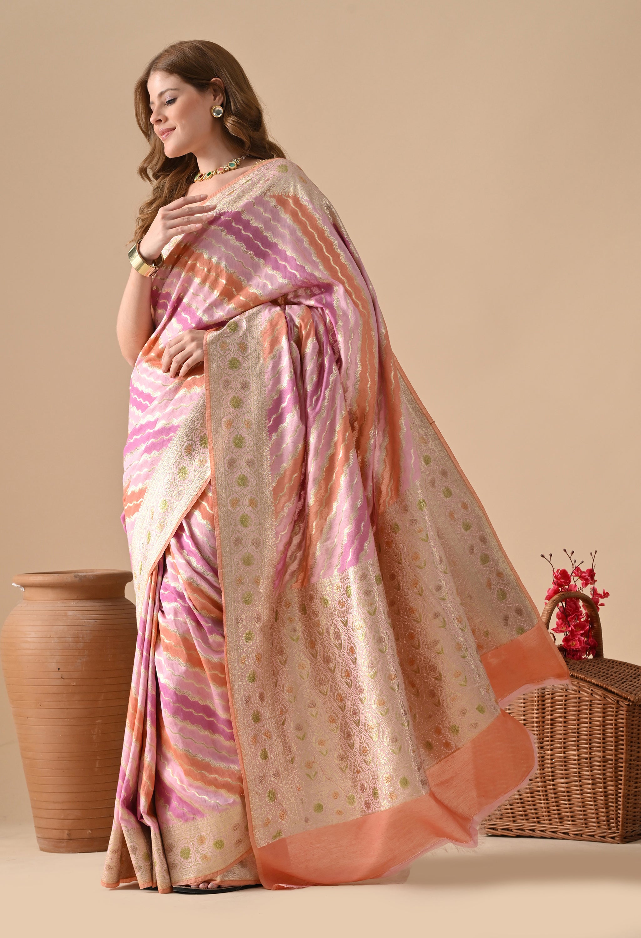 A side-angle studio shot of a woman in a The Weavers Shop by Raavi Banarasi silk saree, featuring the Rangkat technique with diagonal wavy stripes in shades of pink, lavender, and orange. The saree is adorned with a rich gold zari border and a matching pallu with traditional floral motifs and tassel detailing. She is styled with a gold blouse, a green gemstone choker, and a neat bun, positioned between a large clay pot and a wicker basket of red flowers.