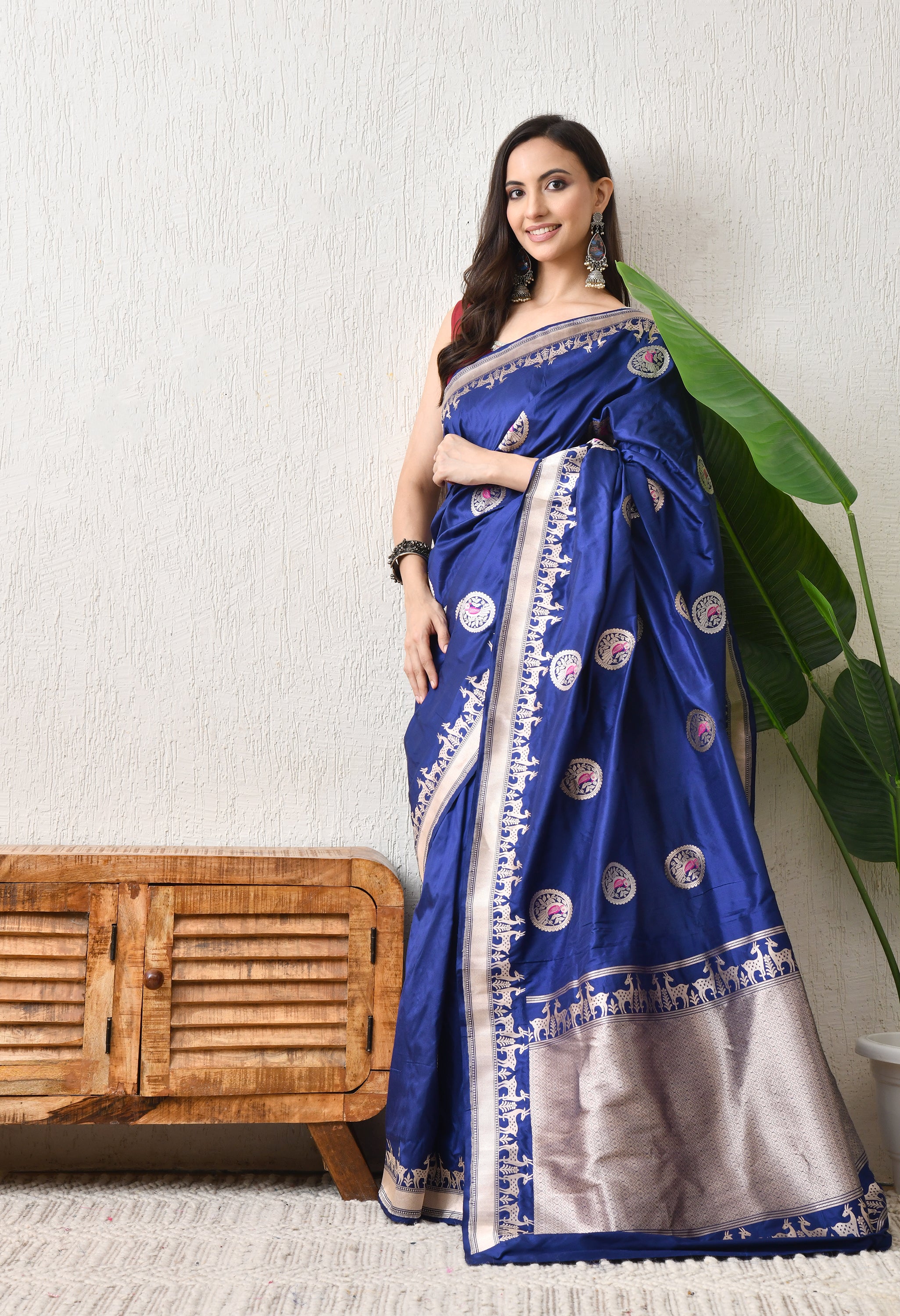 A side-frontal view of a woman posing in the royal blue Hand Woven Katan Silk saree from The Weavers Shop by Raavi. This angle highlights the rich luster of the Silk Mark Certified fabric and the repeating deer motifs of the Shikarga border. She is styled with traditional silver jewelry, showcasing the saree's suitability for royal festive or wedding wear.