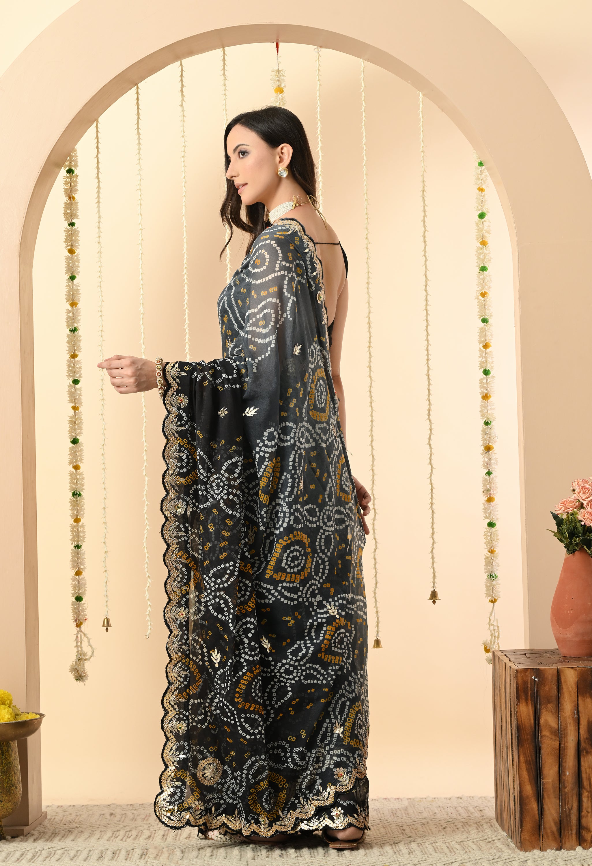 A woman is pictured from the back, showcasing the full drape and intricate design of a black georgette saree from The Weavers Shop by Raavi. This handcrafted Rajasthani Bandhani saree, perfect for a festive event or a classy evening party, features traditional white and yellow tie-dye patterns and is detailed with a gold Gota Patti scalloped border. She is styled with a sleeveless black blouse and a white pearl choker, standing against a neutral-toned archway decorated with jasmine garlands.