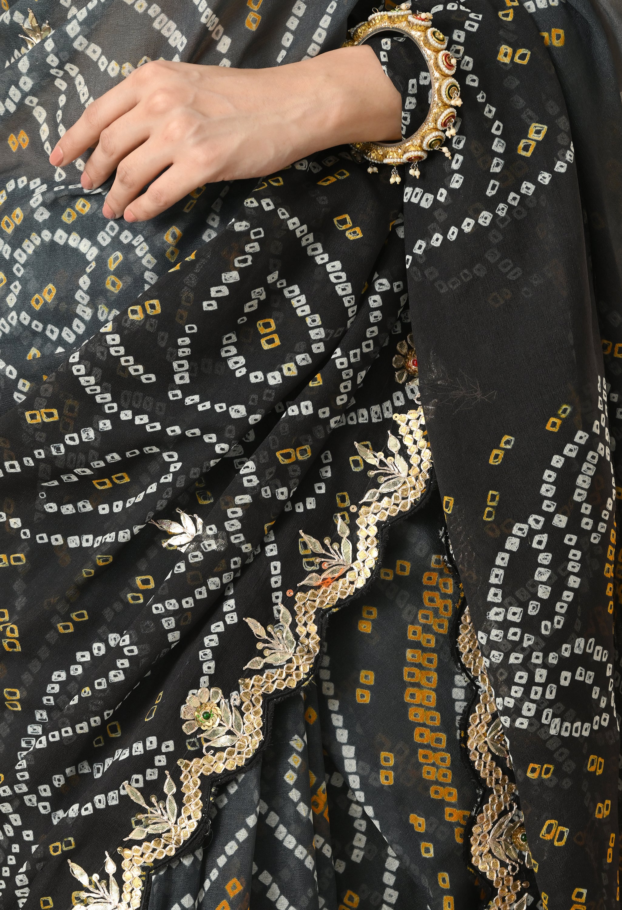 A vertical close-up shot focusing on the intricate textures and craftsmanship of a black georgette saree from The Weavers Shop by Raavi. This handcrafted Rajasthani Bandhani saree features traditional white and mustard-yellow tie-dye patterns alongside a scalloped border with gold Gota Patti embroidery and floral leaf motifs. The woman's hand is visible, adorned with a heavy traditional gold bangle featuring colorful stone inlay and hanging pearl droplets.