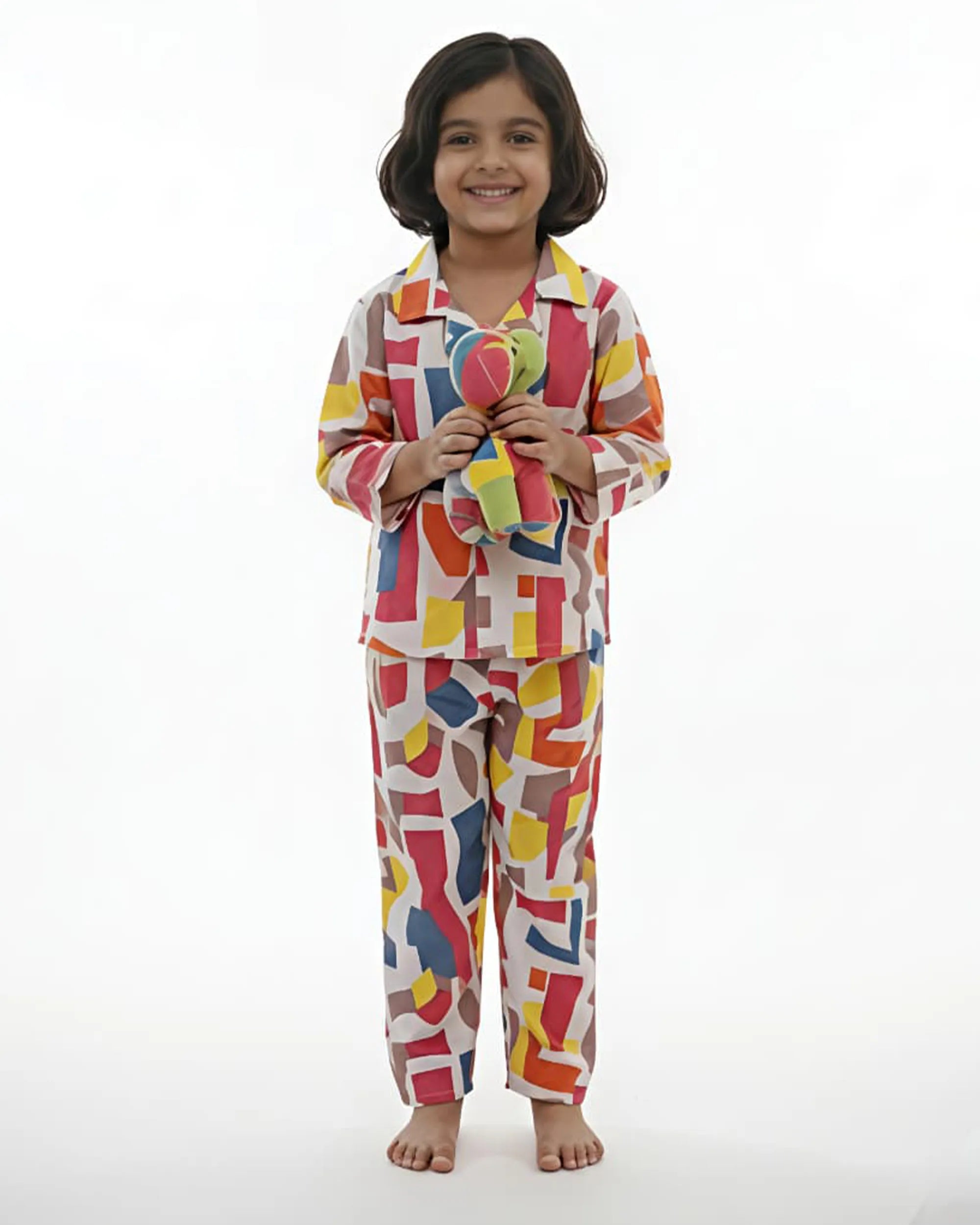 A young girl standing against a white background, wearing a white long-sleeved button-up pajama set with a colorful, multi-colored geometric square print