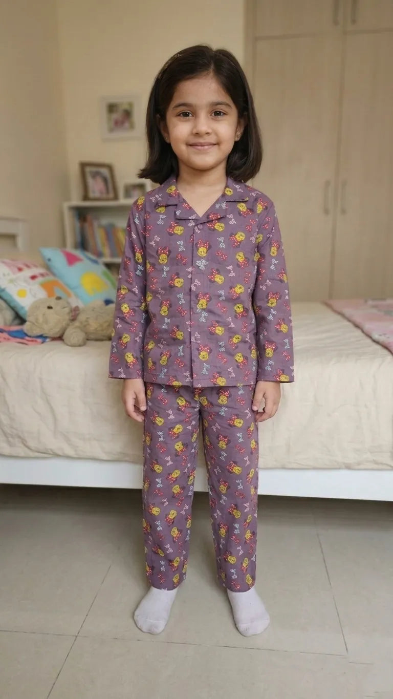 A young girl smiling while wearing a mauve purple cotton night suit with a cartoon character and bow print.
