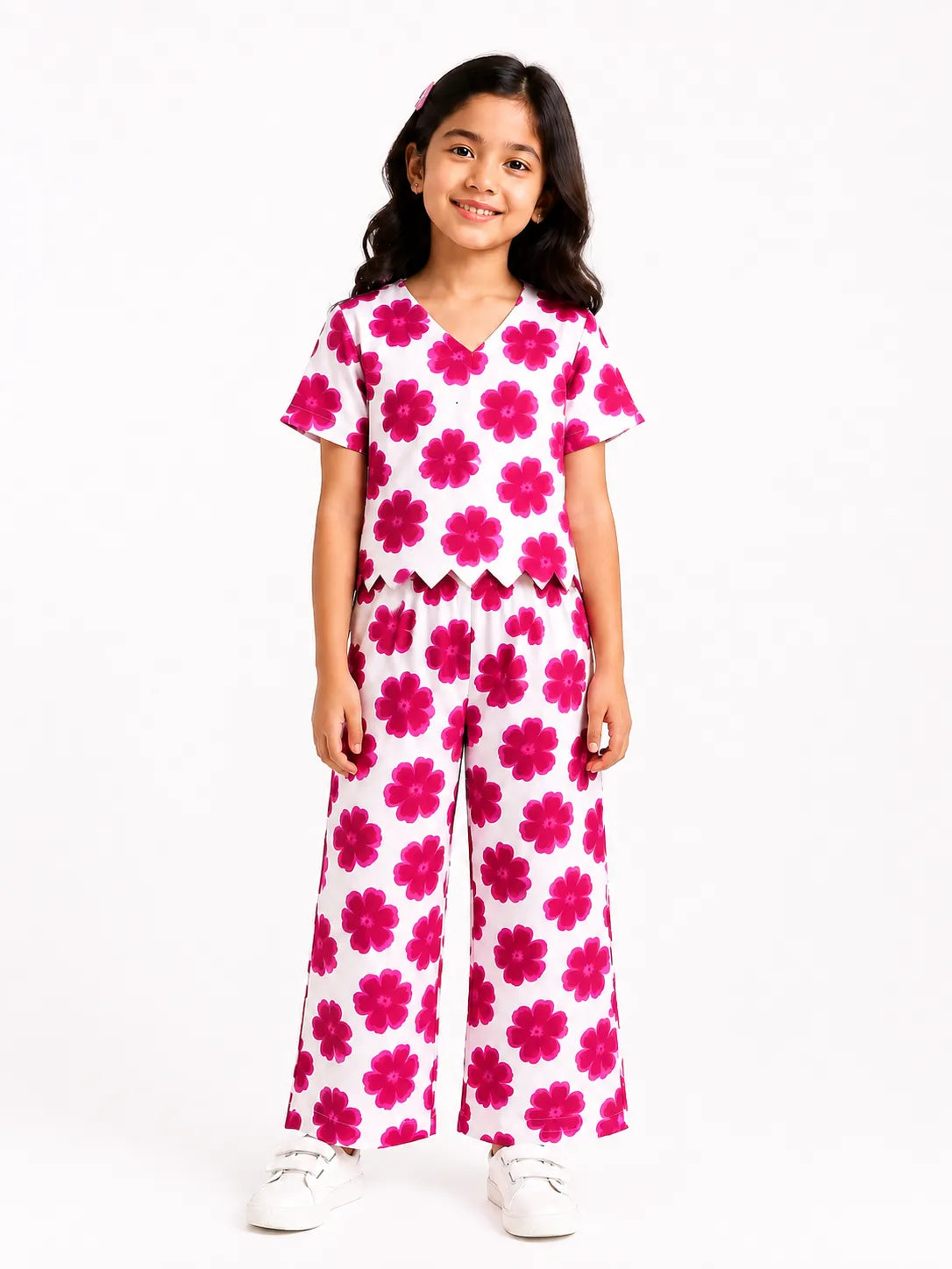 Young girl smiling in a white two-piece co-ord set featuring a bold pink floral pattern, a V-neck scallop-hem top, and matching wide-leg pants.
