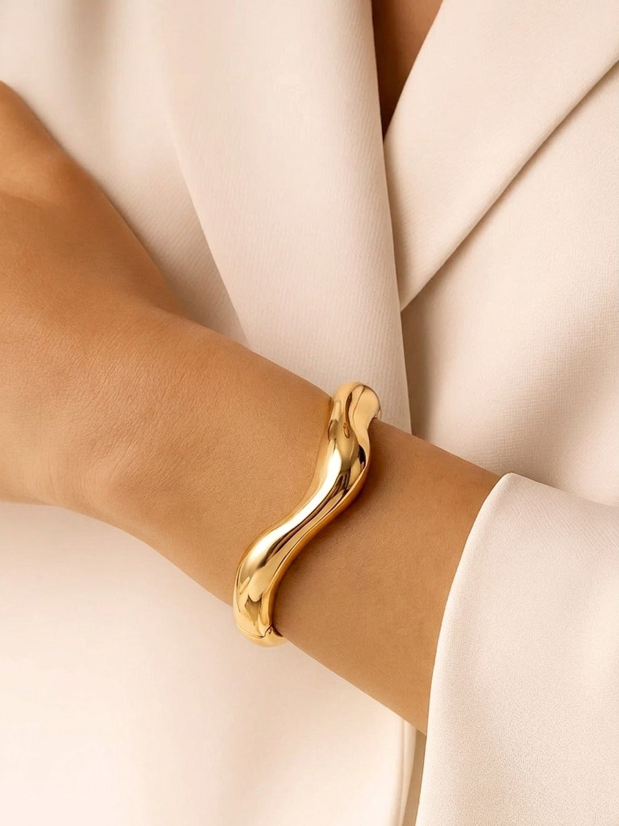 A high-shine gold bangle featuring a sculptural wave design and a side hinge, displayed upright on a light surface.
