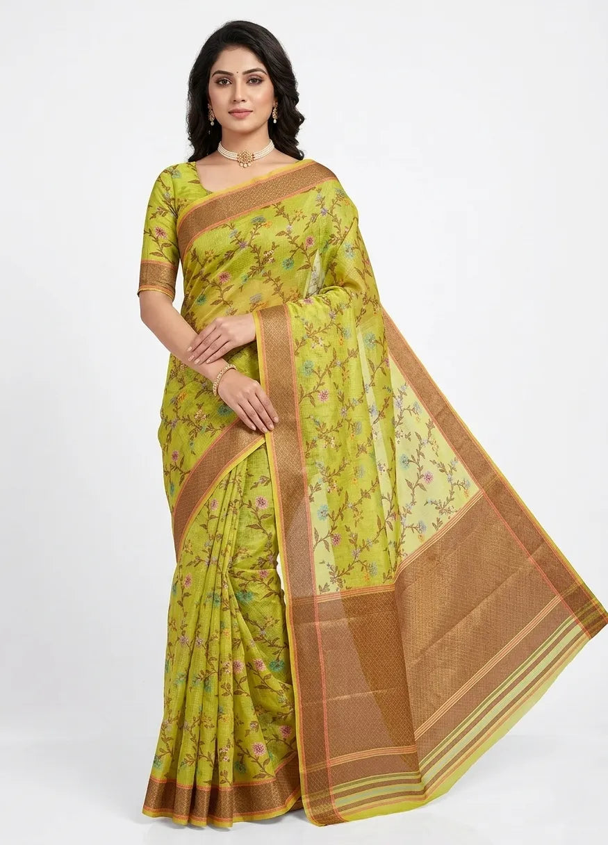 An Indian woman modeling a vibrant lime green organza saree featuring small, multi-colored floral prints. The saree is finished with a wide, bronze-colored zari border. She is wearing a matching printed blouse, a pearl necklace, and gold earrings, standing against a plain white background.
