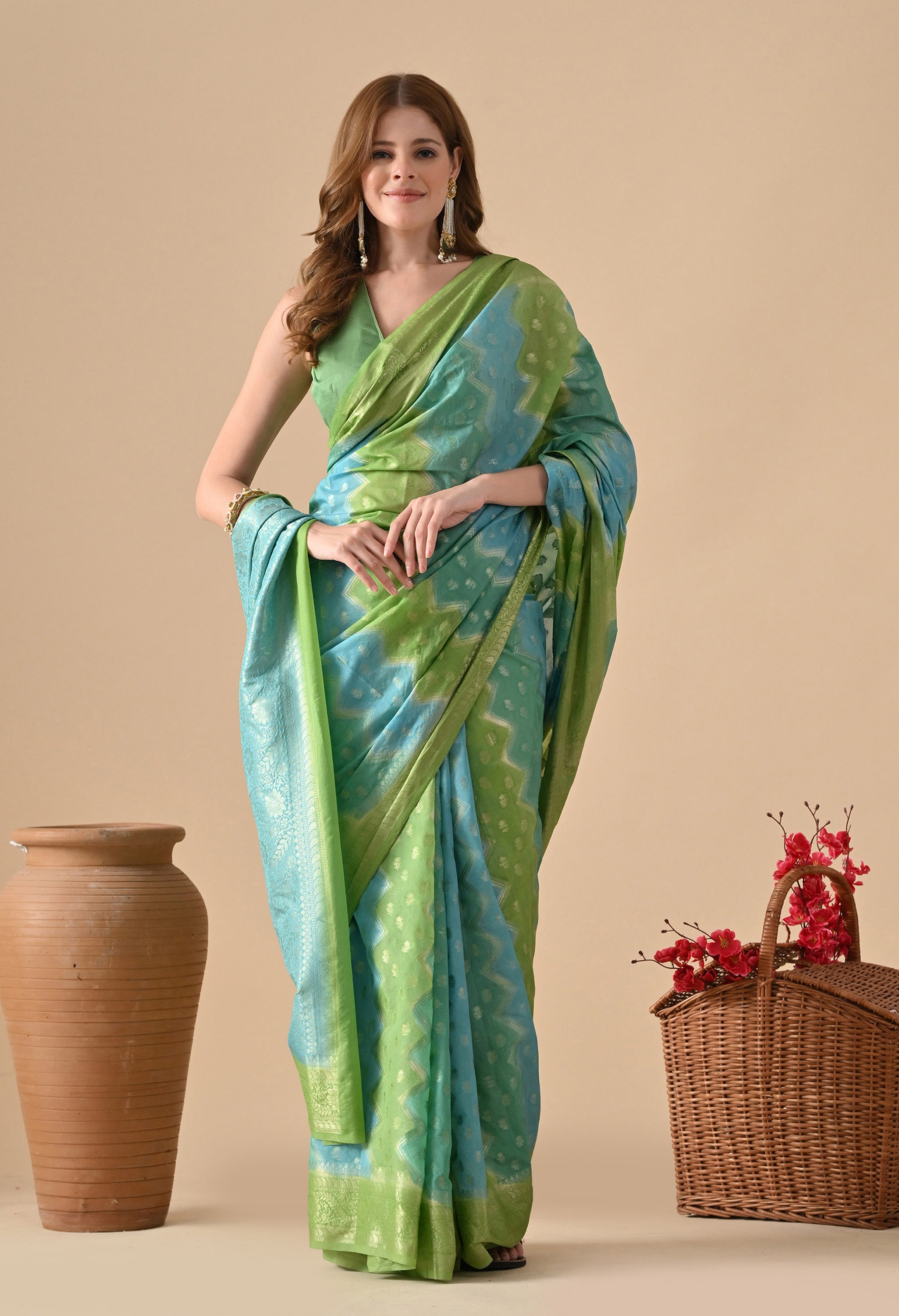 A woman models a Banarasi Rangkat saree from The Weavers Shop, featuring a vibrant diagonal "Rangkat" pattern in alternating shades of seafoam green and sky blue. The saree is adorned with intricate gold zari buttas (small floral motifs) and a traditional zari border. She pairs it with a sleeveless lime green blouse, long statement earrings, and a gold bangle, standing against a neutral beige background next to a tall clay pot and a wicker basket of pink flowers.