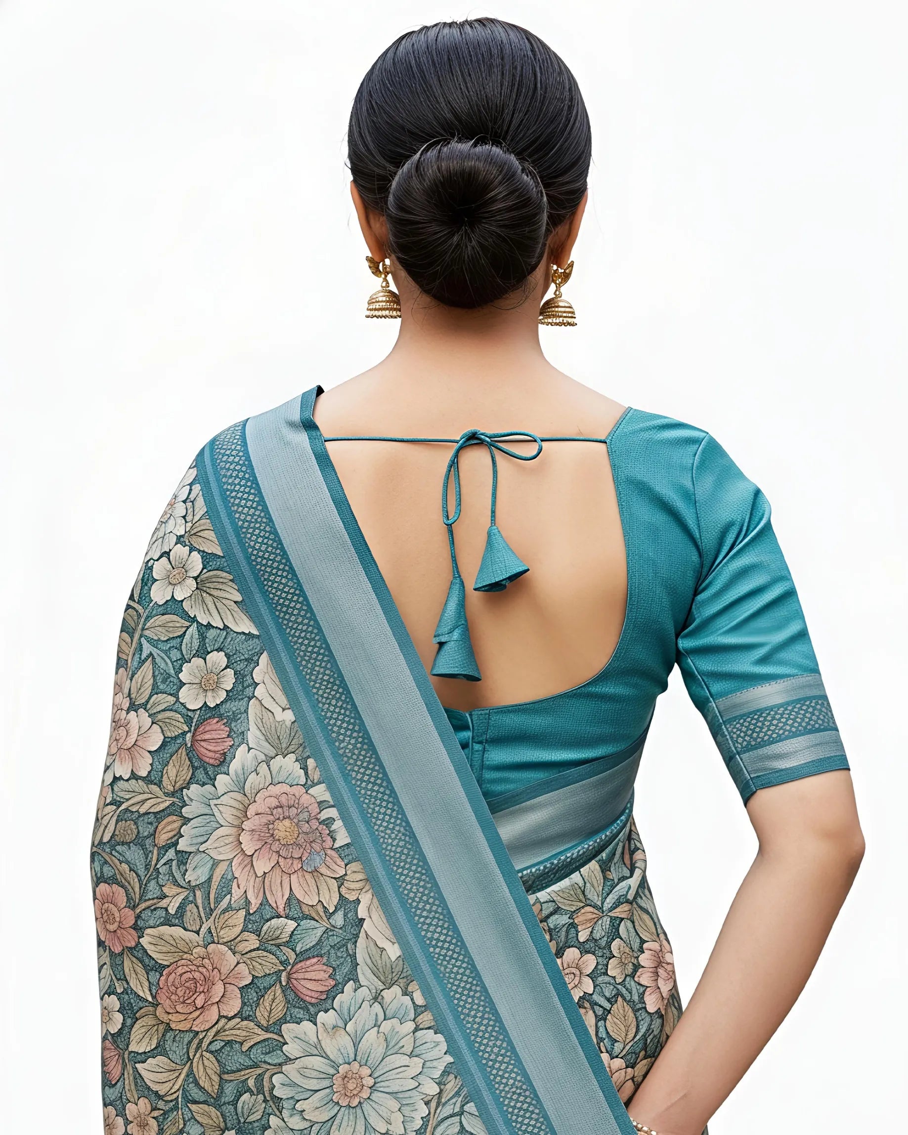 Close-up back view of a teal green floral cotton saree. The matching teal blouse features a deep U-back design with delicate tie-up tassels (dori). The saree's pallu is shown with intricate geometric border patterns and small fringe tassels at the end.