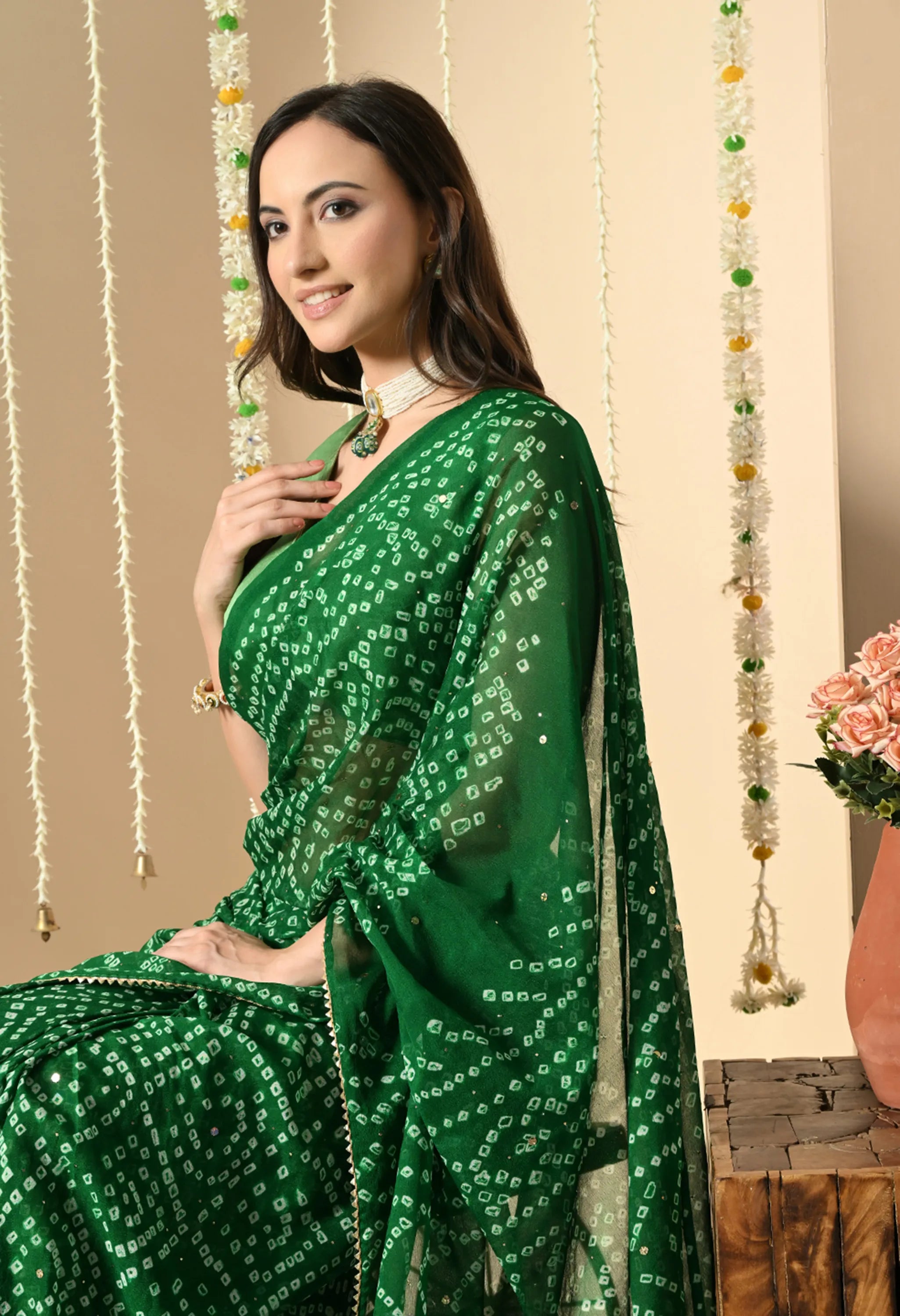 A close-up of a woman smiling softly while wearing a vibrant green georgette sari from The Weavers Shop by Raavi. The sari features a traditional white Bandhani pattern with a rich gold zari border, ideal for a festive event. She is adorned with a pearl choker and jhumka earrings, posed against a festive backdrop of orange and yellow marigold garlands.