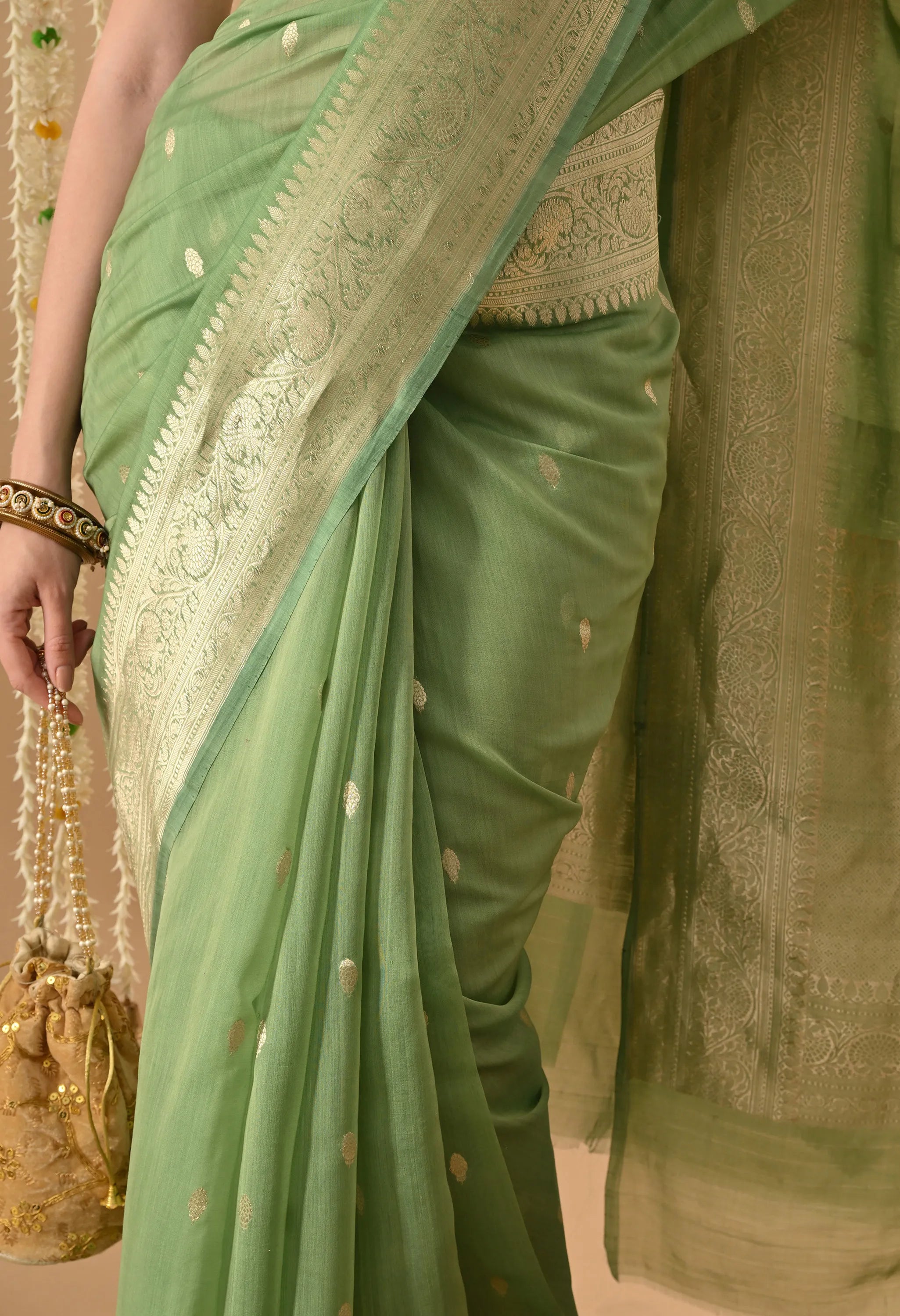 A detailed mid-section view of a Silk Mark Certified mint green pure silk saree from The Weavers Shop by Raavi. The image highlights the hand-woven Karuwa Weave, featuring small gold zari work motifs meticulously woven onto the soft, luxurious pure silk fabric. The focus is on the elegantly draped pleats, which showcase the smooth fall and rich texture of the silk. The model holds a traditional gold embellished potli bag with pearl strings, and the broad gold zari border with its dense floral-vine pattern 