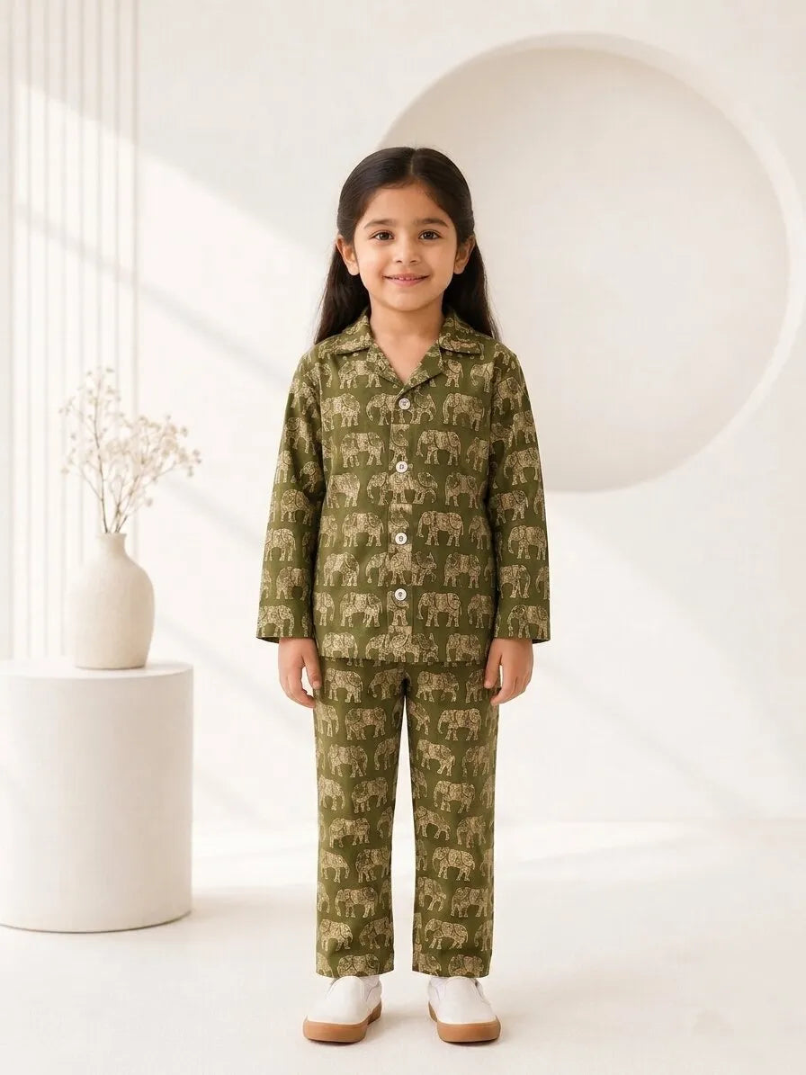 A young girl smiling while wearing a green cotton pajama set featuring an intricate olive-green elephant pattern. She is standing in a brightly lit room with a blurred background of toys and books.