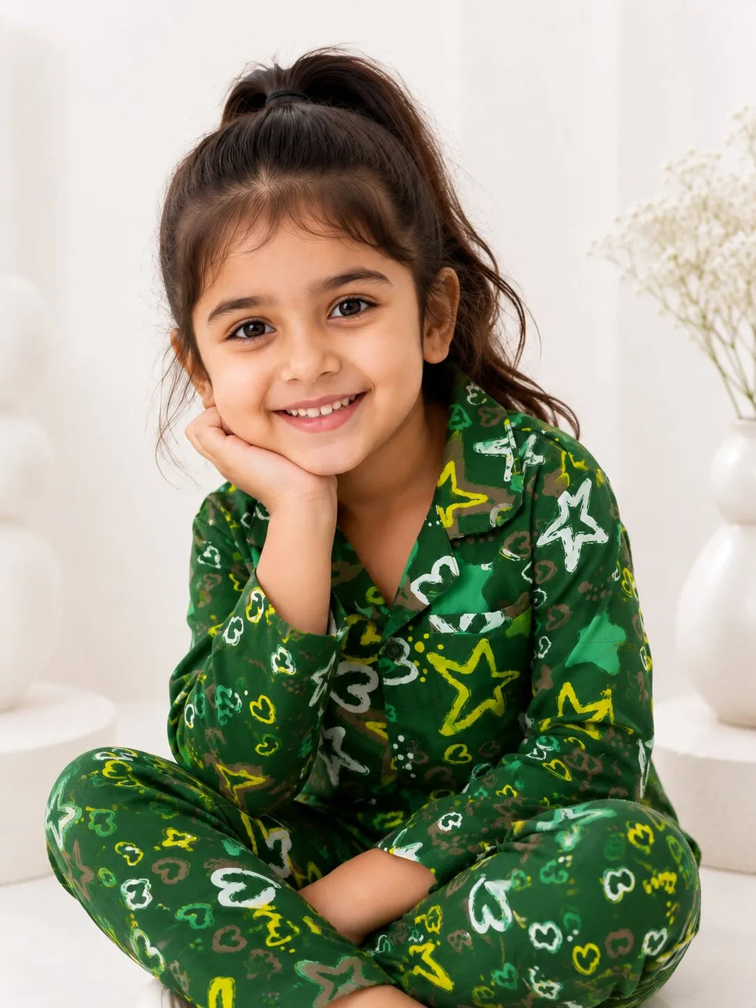 
A young girl with long, wavy dark hair smiling brightly while holding the collar of her pajama top. She is wearing a white long-sleeve pajama shirt featuring a vibrant, colorful repeating pattern of hot air balloons in shades of yellow, purple, pink, and orange, set against a plain white background.