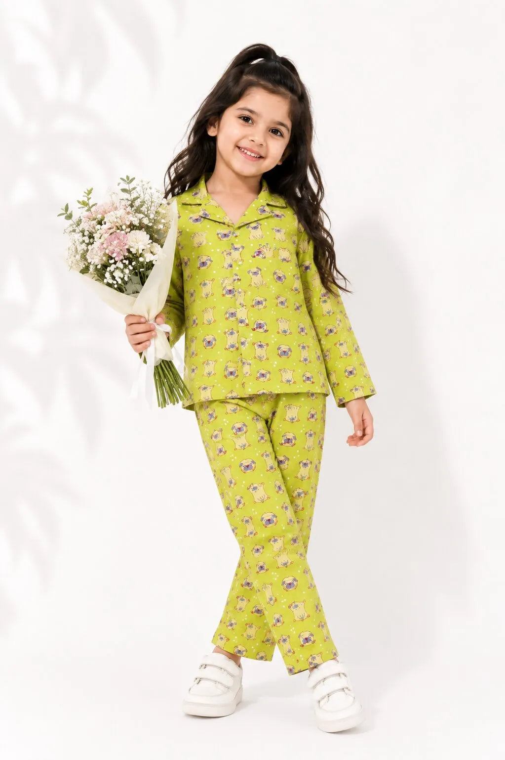 A young girl smiling in a lime green cotton night suit with a pug dog print, standing in a cozy bedroom.