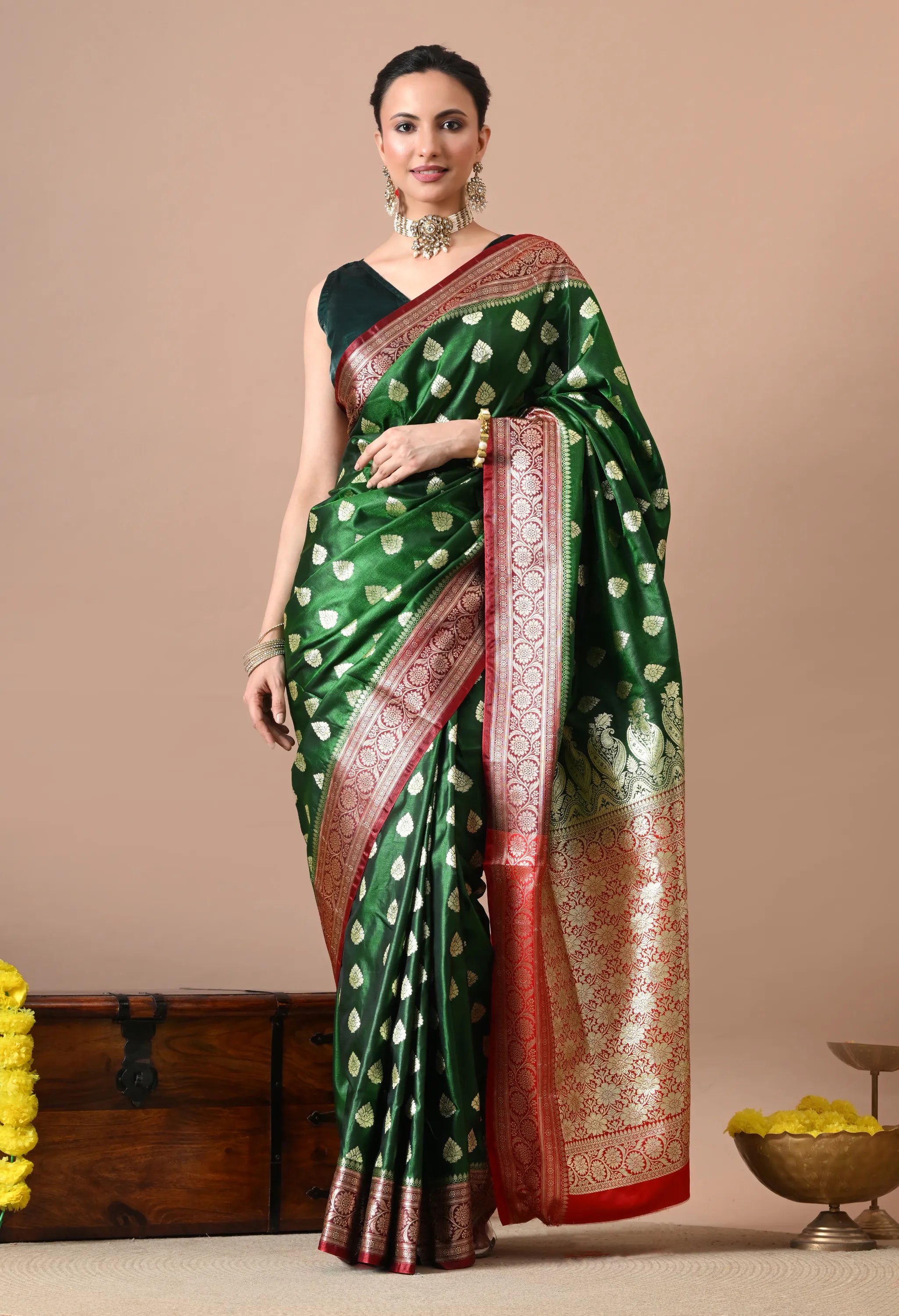Woman modeling a dark green silk-blend saree with gold zari leaf patterns and a thick red-and-gold traditional border.
