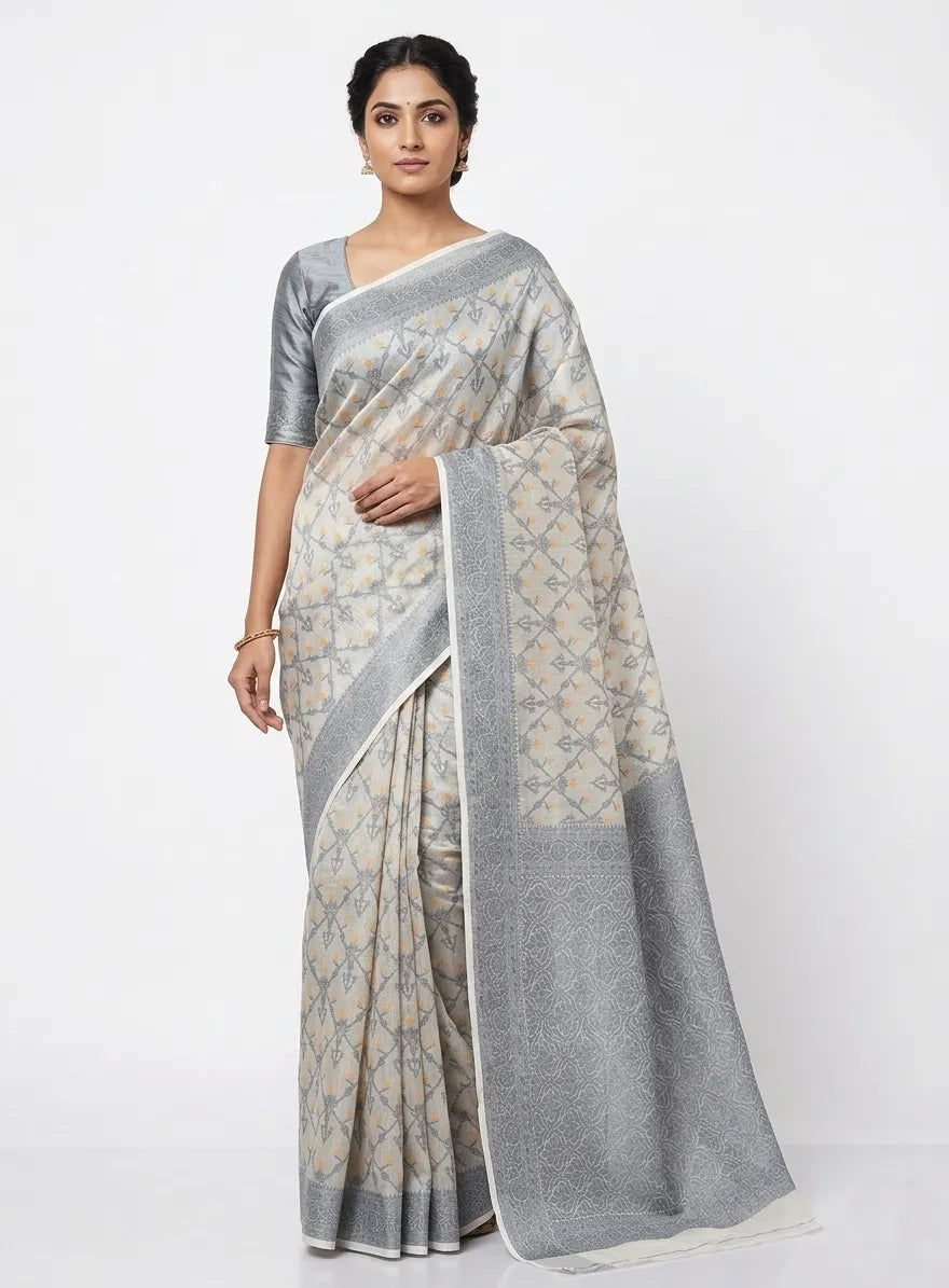 An Indian woman modeling a grey and off-white silk blend saree with a diamond-patterned woven body and a wide silver zari border. She is wearing a solid grey half-sleeved blouse and gold jhumkas against a plain white background.

