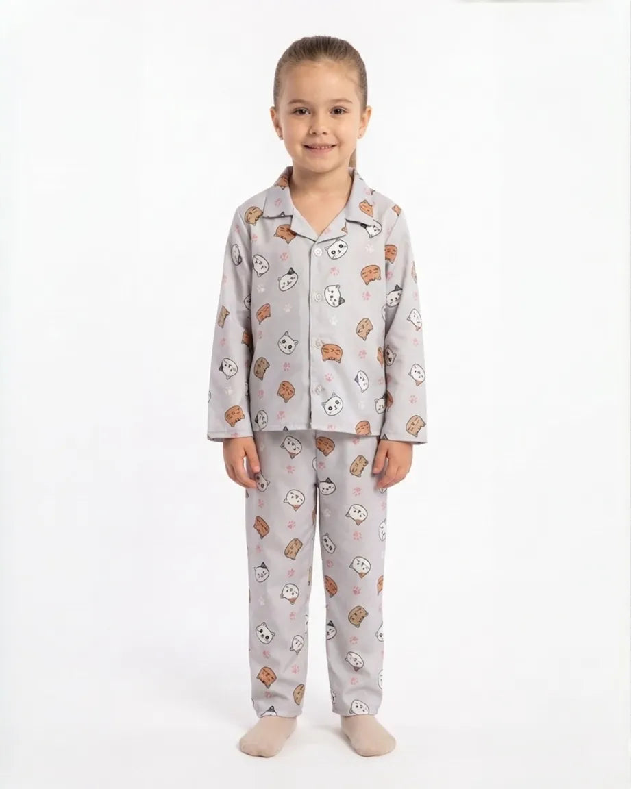 A young girl smiling against a white background, wearing a light grey satin night suit with a playful all-over cat and panda print. The two-piece pajama set features a long-sleeved button-down shirt with a collar and matching full-length trousers.
